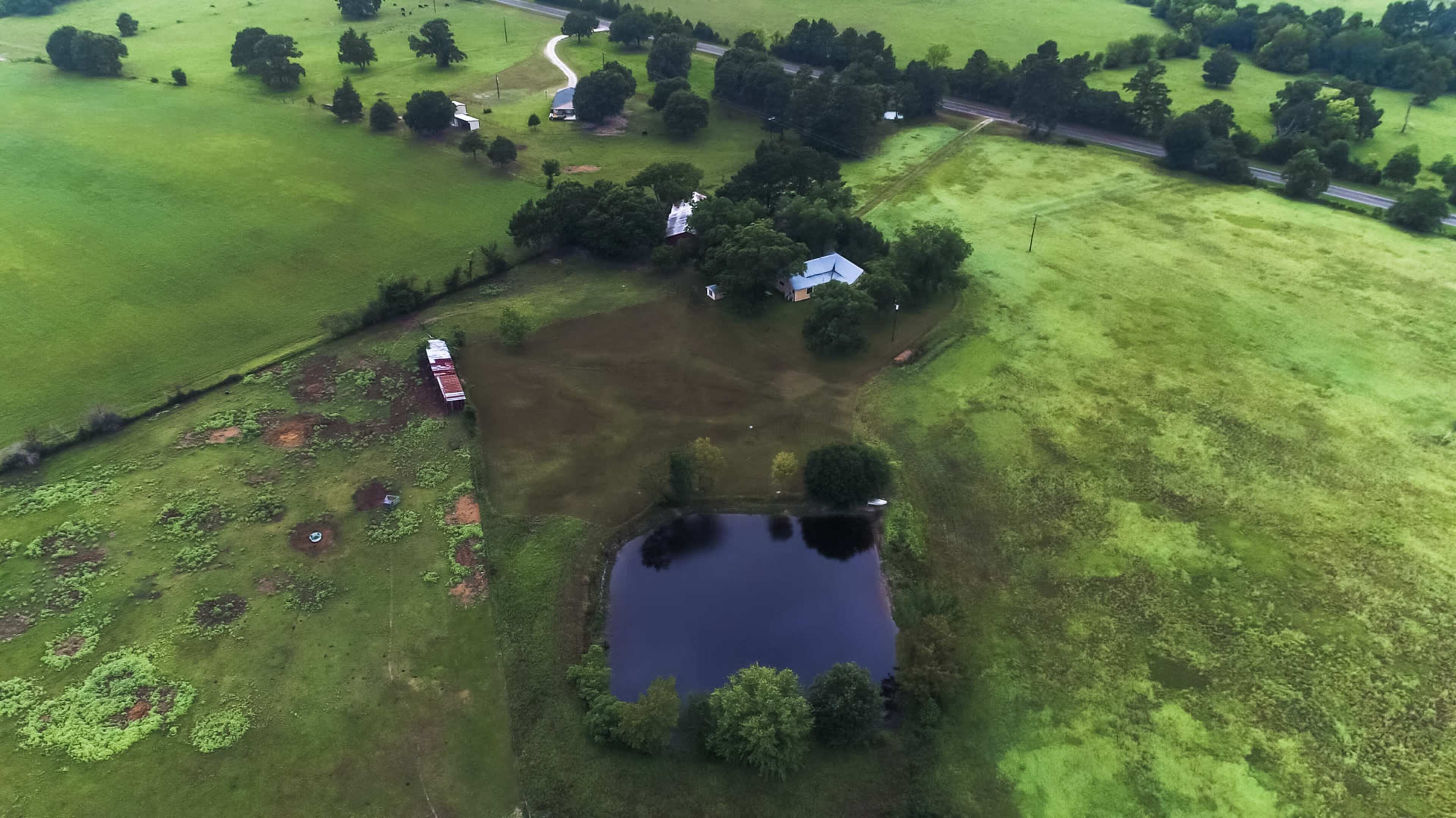 The aerial view shows a rural landscape with a pond, scattered trees, a house near the water, and green fields.
