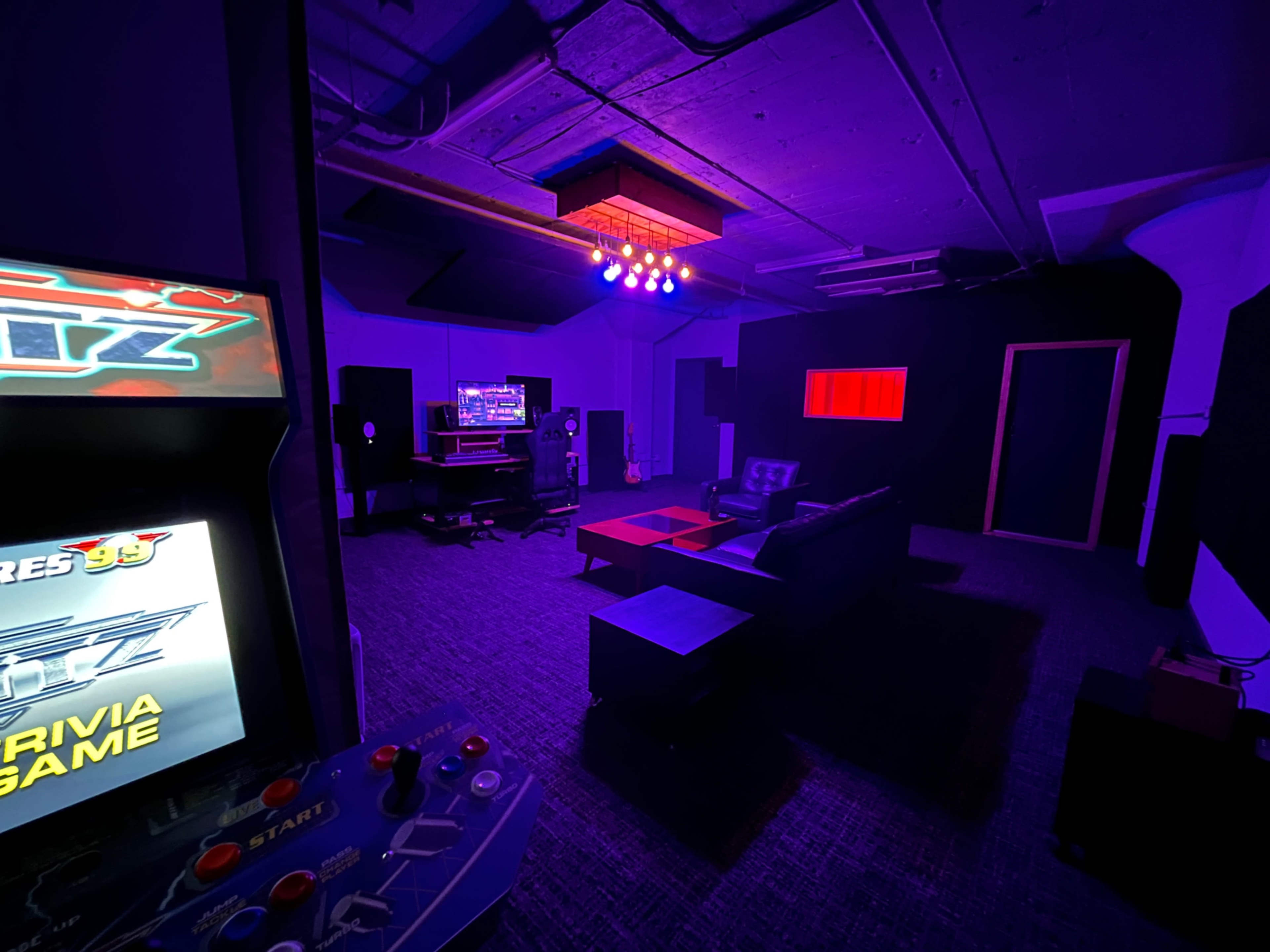 The image shows a dimly lit room featuring a retro arcade game machine, a black sofa set, a coffee table, and audio equipment against walls painted in dark colors.