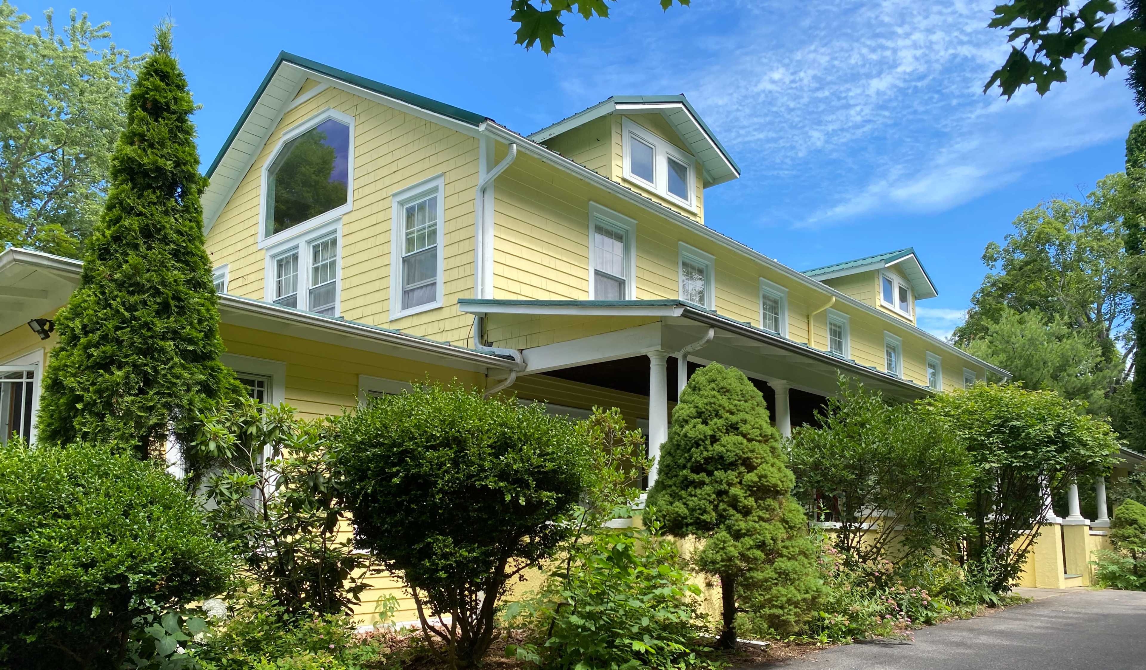 A large yellow house with a peaked roof features multiple windows and is surrounded by neatly trimmed bushes and trees under a clear blue sky.