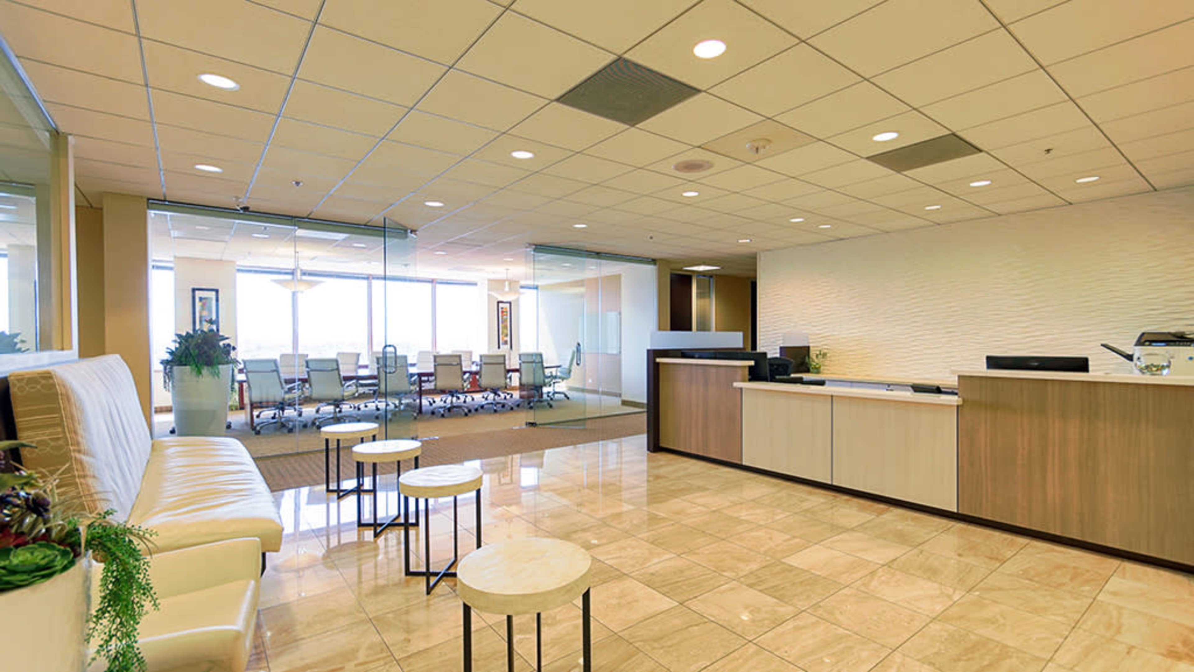 A modern office reception area features a seating space with a white sofa, small round tables, and a reception desk in front of a glass-walled conference room.