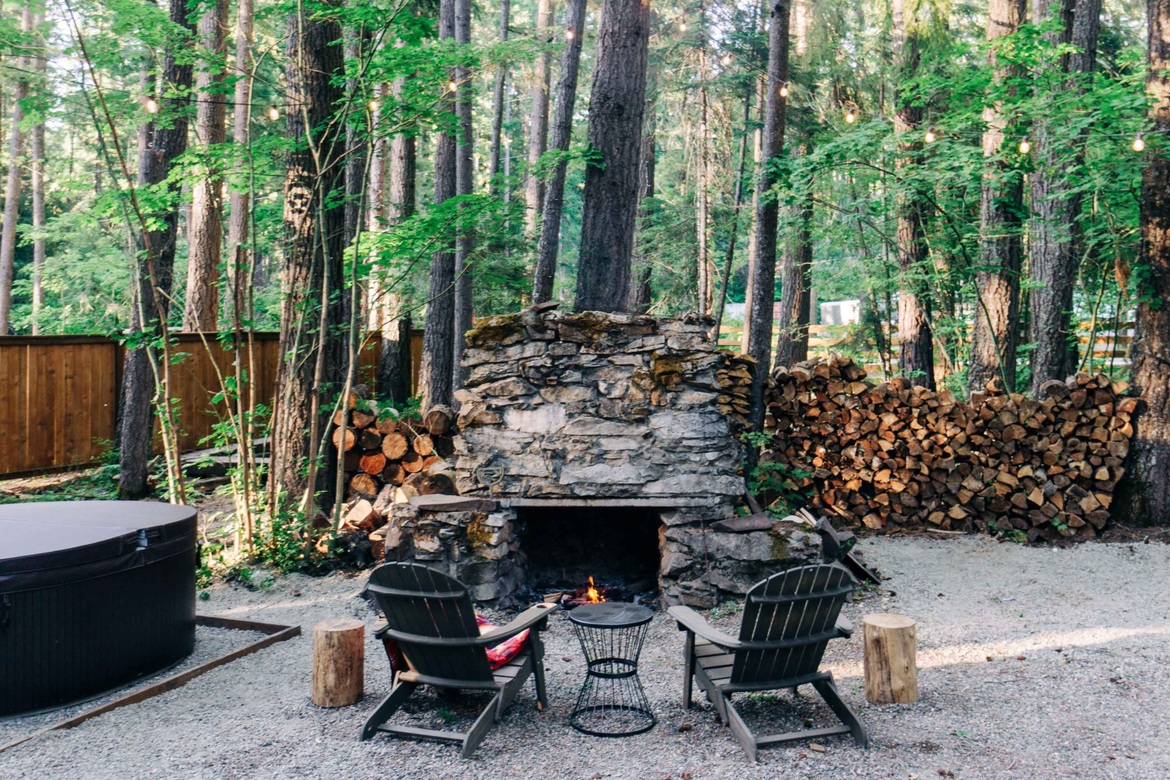 A stone fire pit is surrounded by two chairs in a wooded area with stacked firewood nearby.