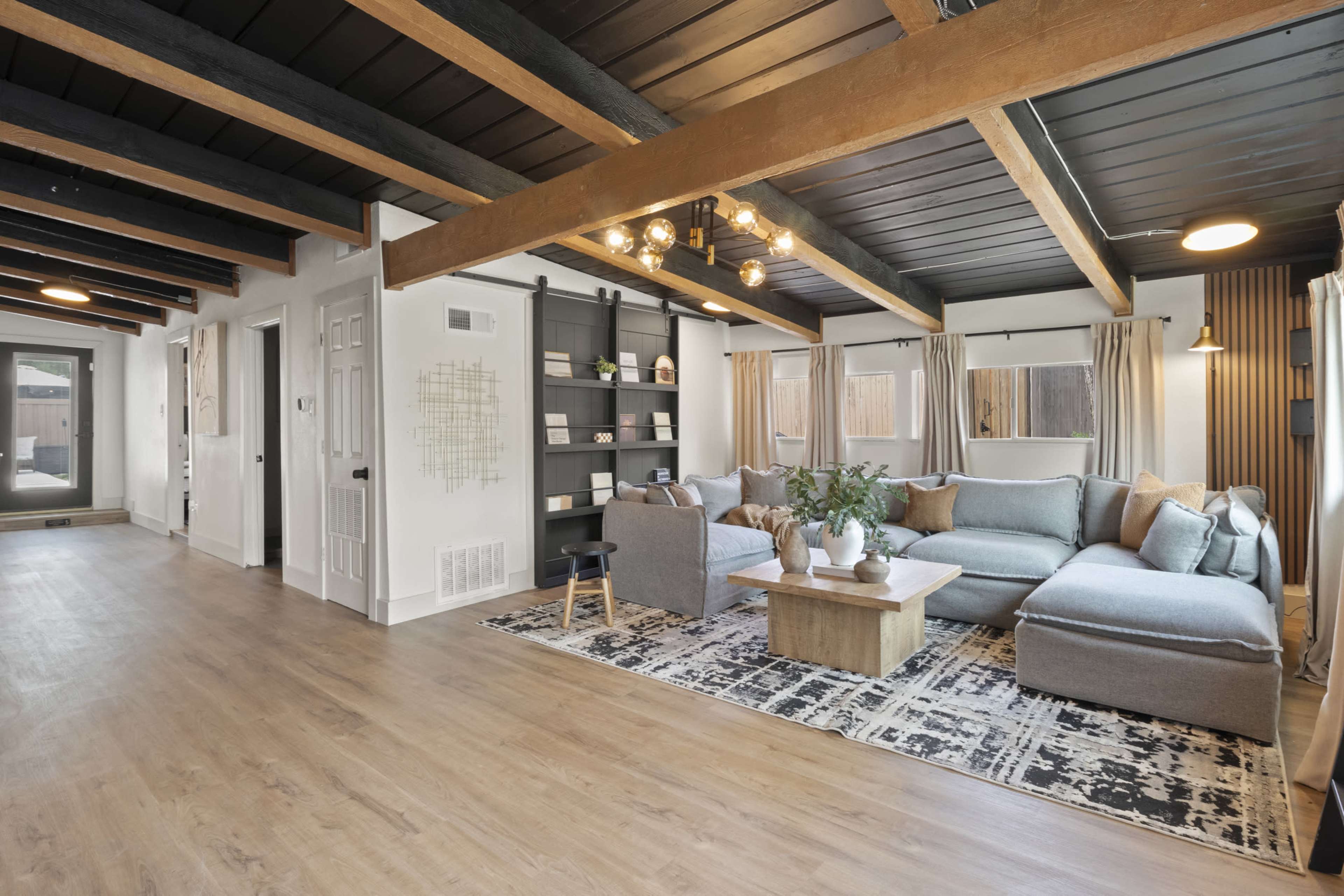 The image shows a modern living room with a sectional sofa, a wooden coffee table, and a bookshelf against a wall, featuring exposed beams on the ceiling and large windows with curtains.