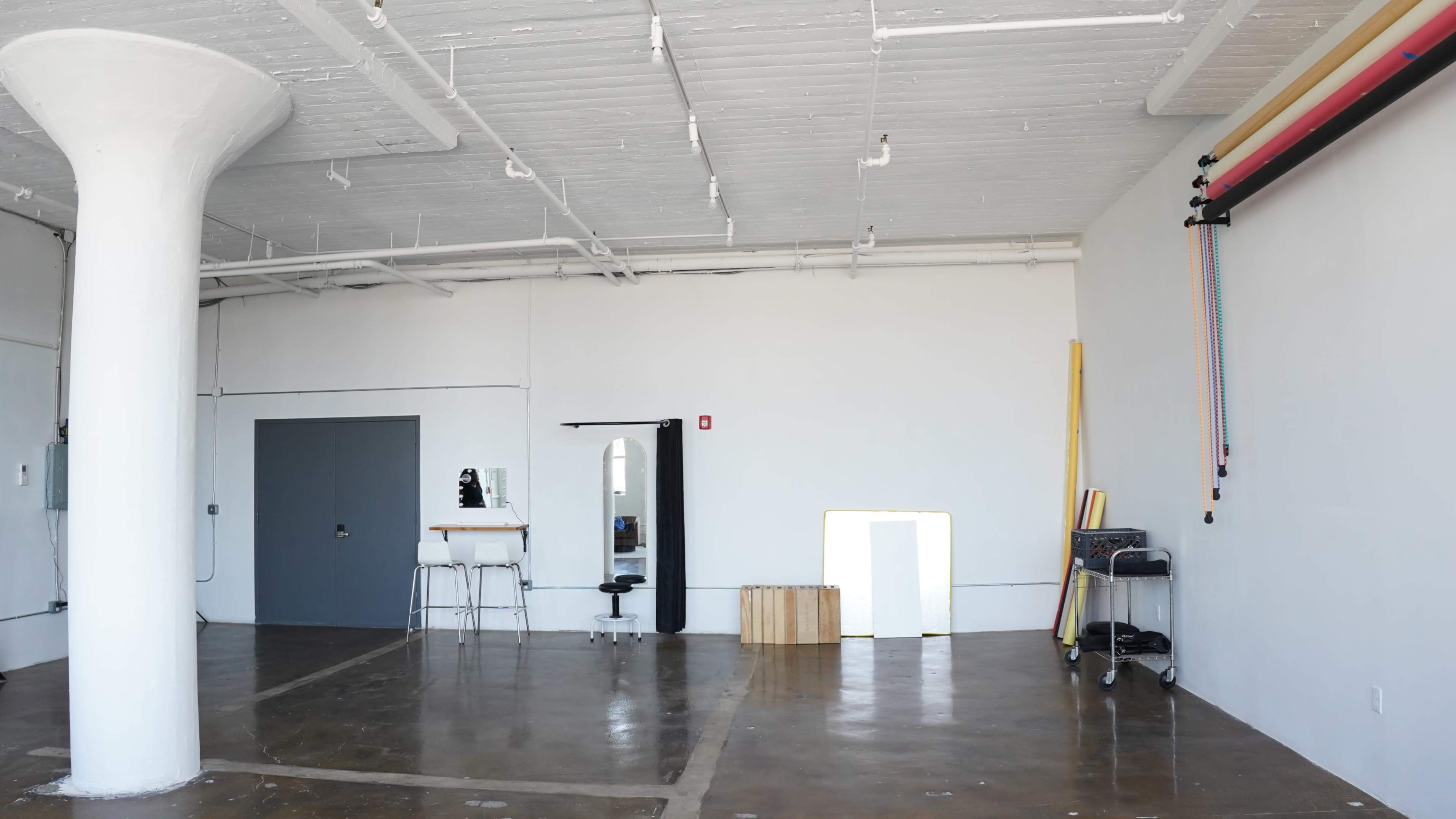 The image shows an empty studio space with concrete flooring, a large mirror, a stool, and some basic furnishings along the walls.