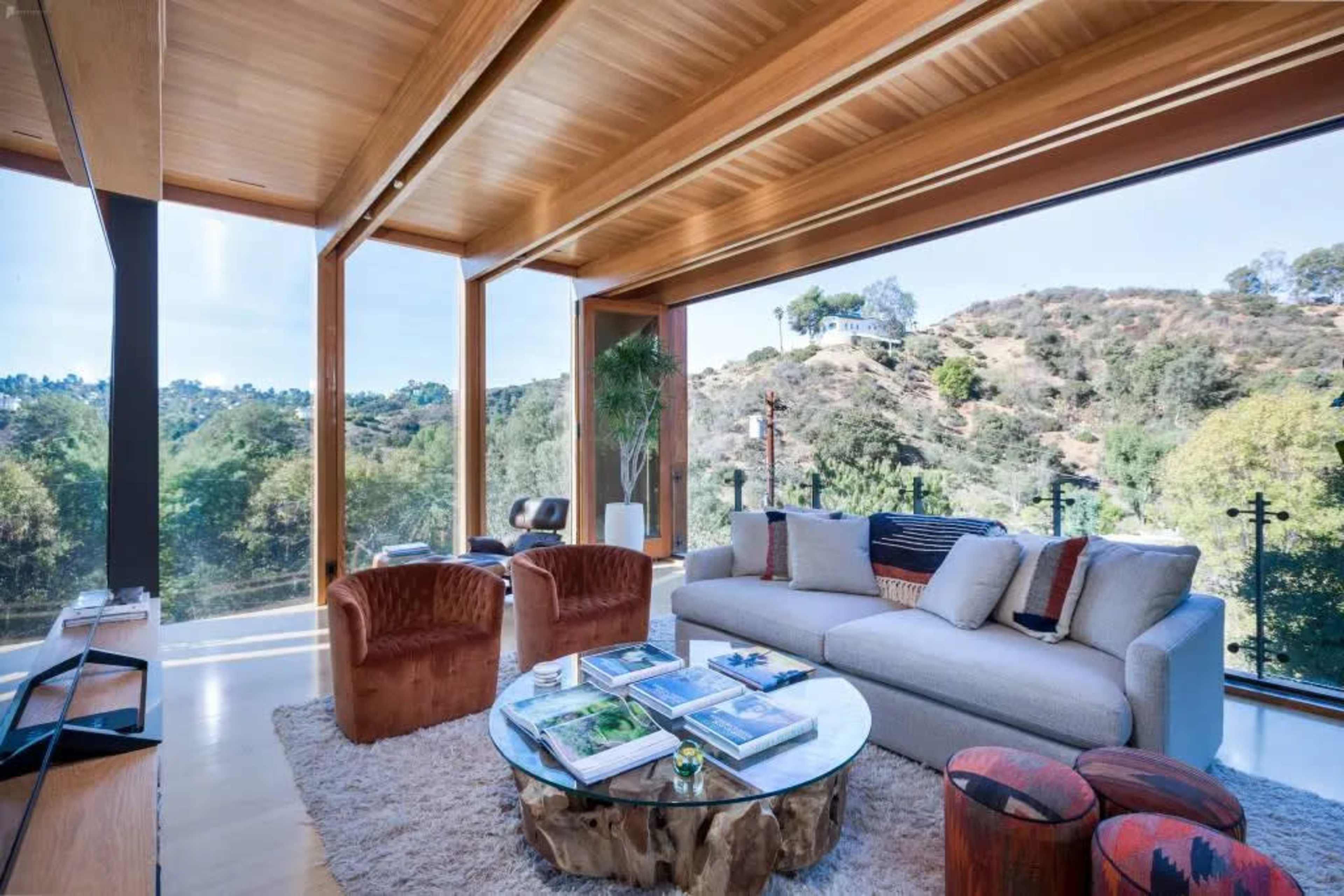 A modern living room features a large window offering a view of a hillside, with a cozy sofa, two armchairs, and a round coffee table arranged on a light-colored rug.