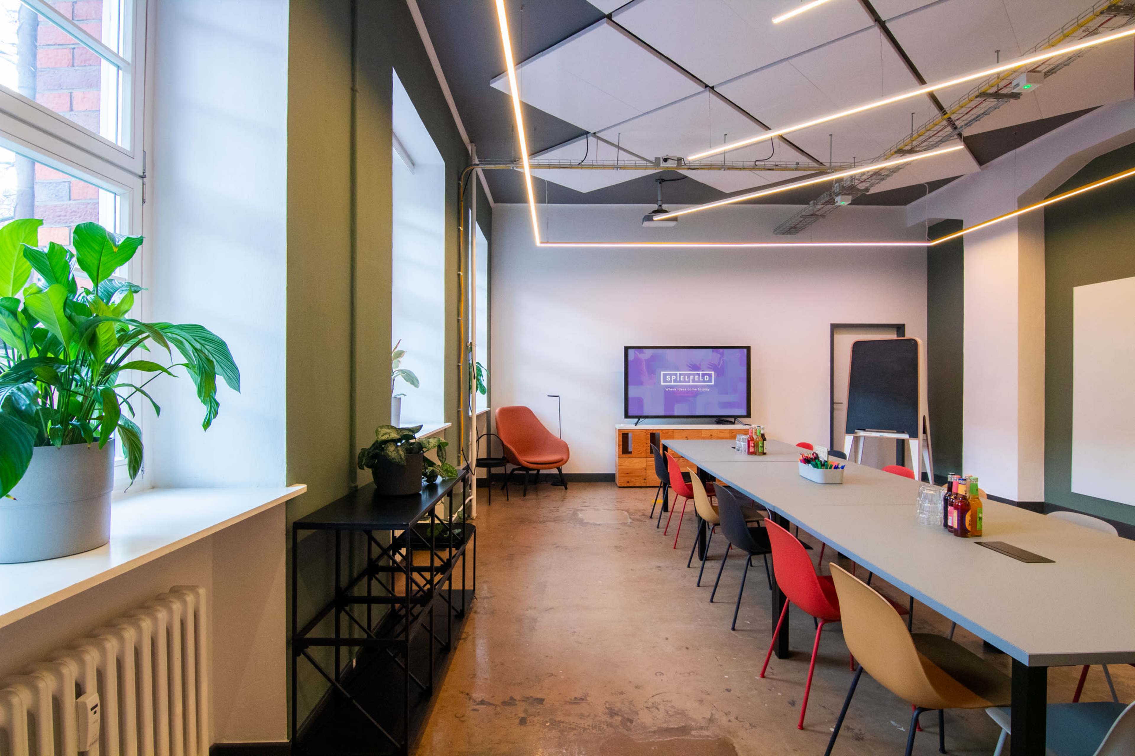 A modern conference room with a long table, colorful chairs, a large screen at one end, and plants near the windows.