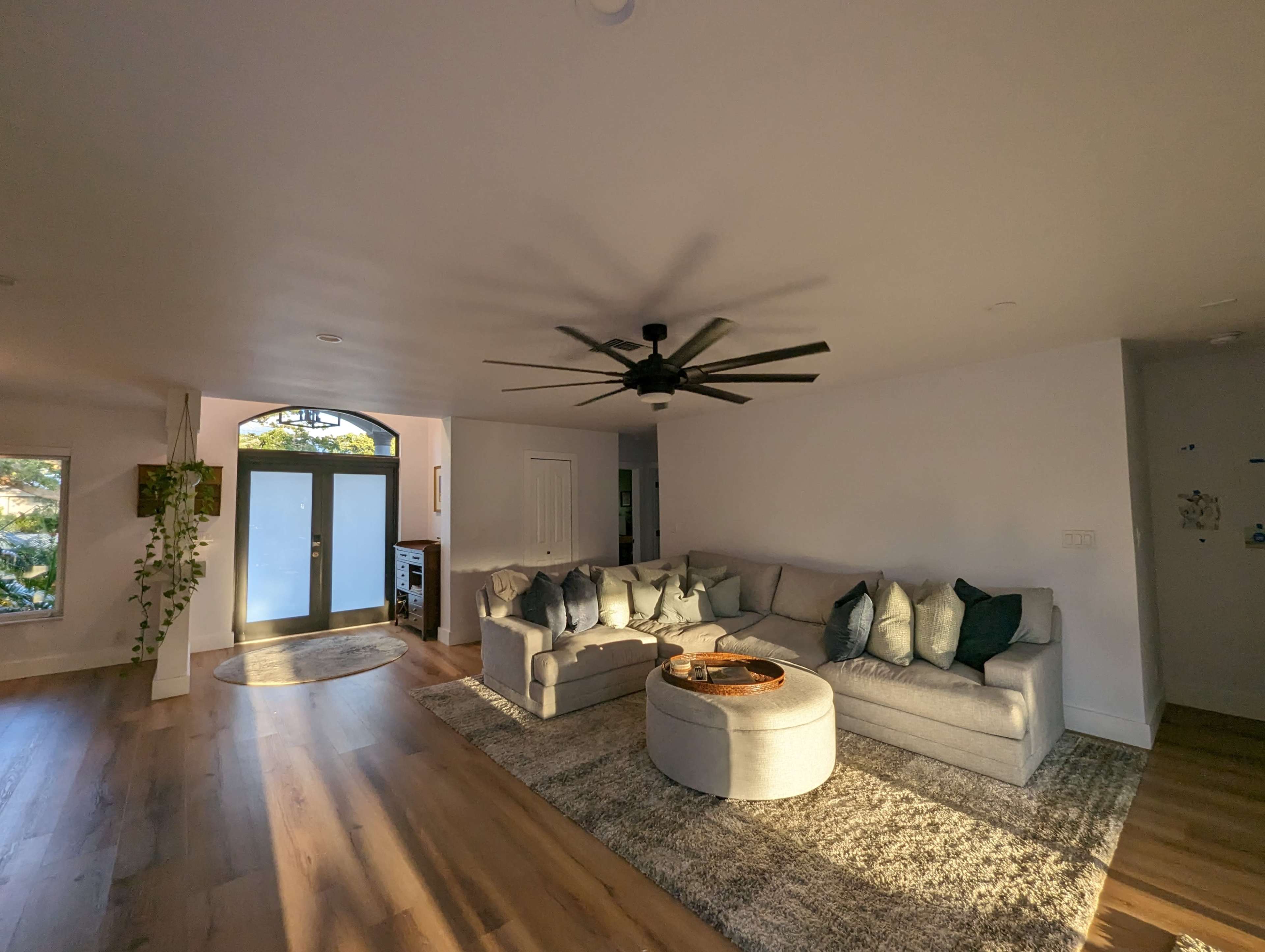 A modern living room with a large gray sectional sofa, a circular coffee table, and a ceiling fan, illuminated by natural light from large windows.