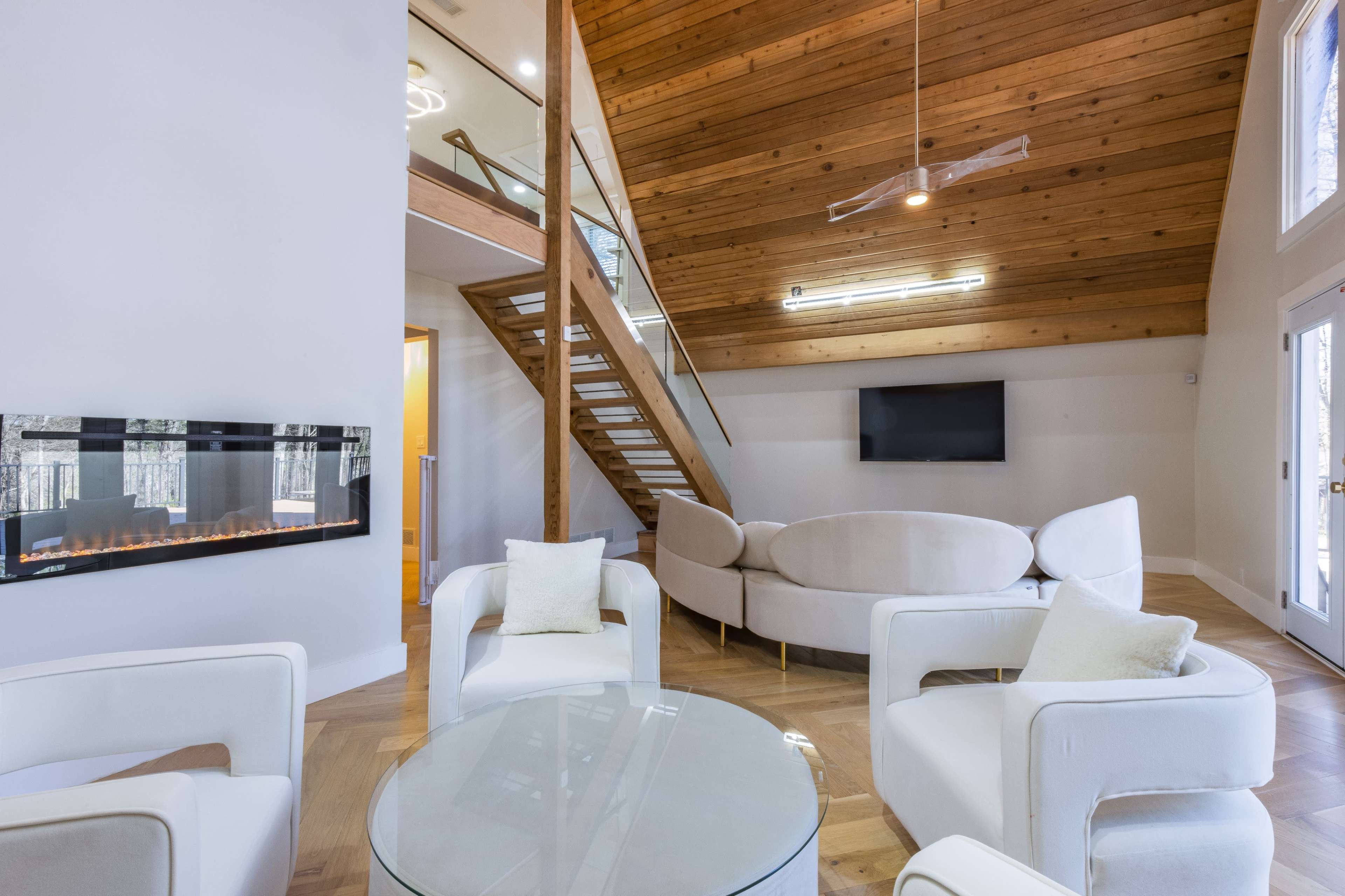 A modern living room features a circular sofa, glass coffee table, and a linear fireplace, with a staircase leading to an upper level.