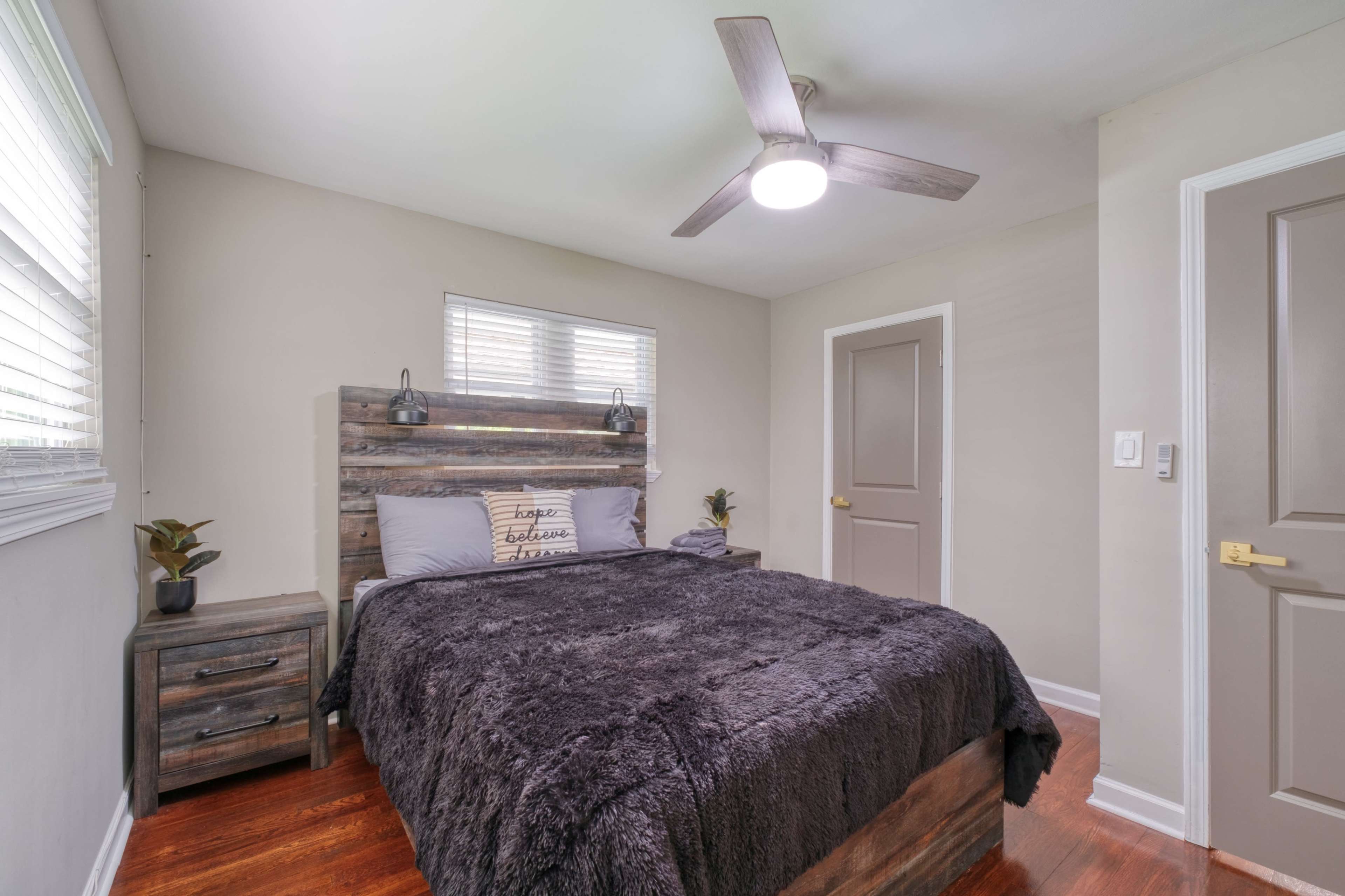 The image shows a cozy bedroom with a wooden bed frame, a nightstand, and a ceiling fan, featuring neutral-colored walls and a window with blinds.