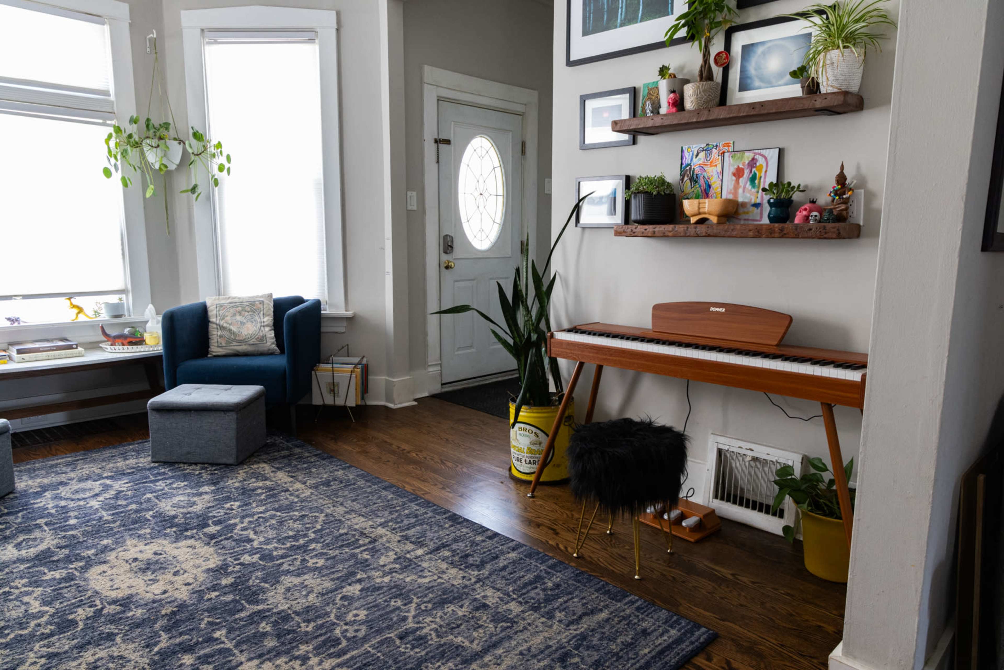 A living room features a blue rug, a piano on a stand, a potted plant, and a shelf with decorative items.
