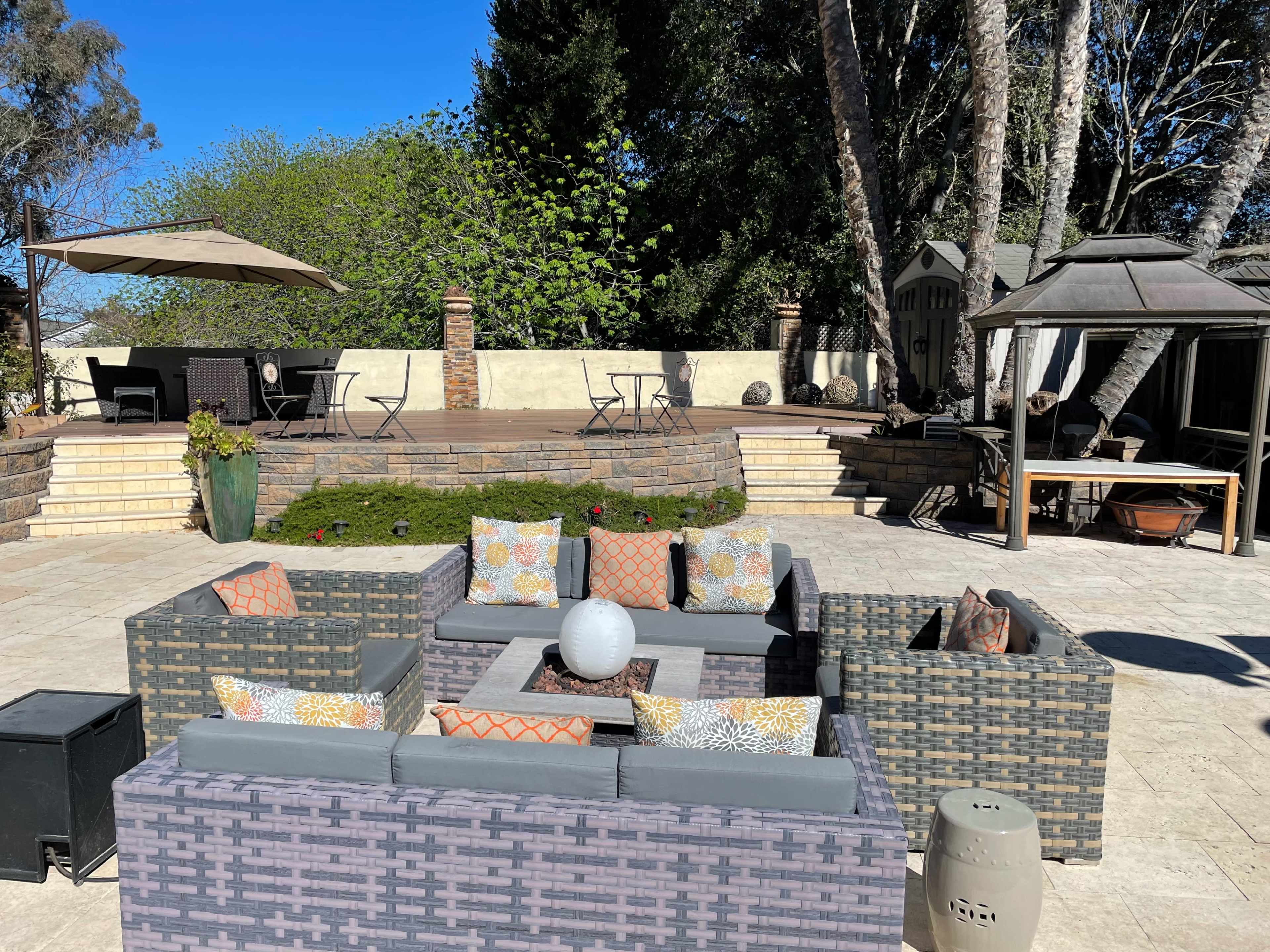 An outdoor seating area with a gray woven furniture set arranged around a central fire pit, surrounded by greenery and a patio.