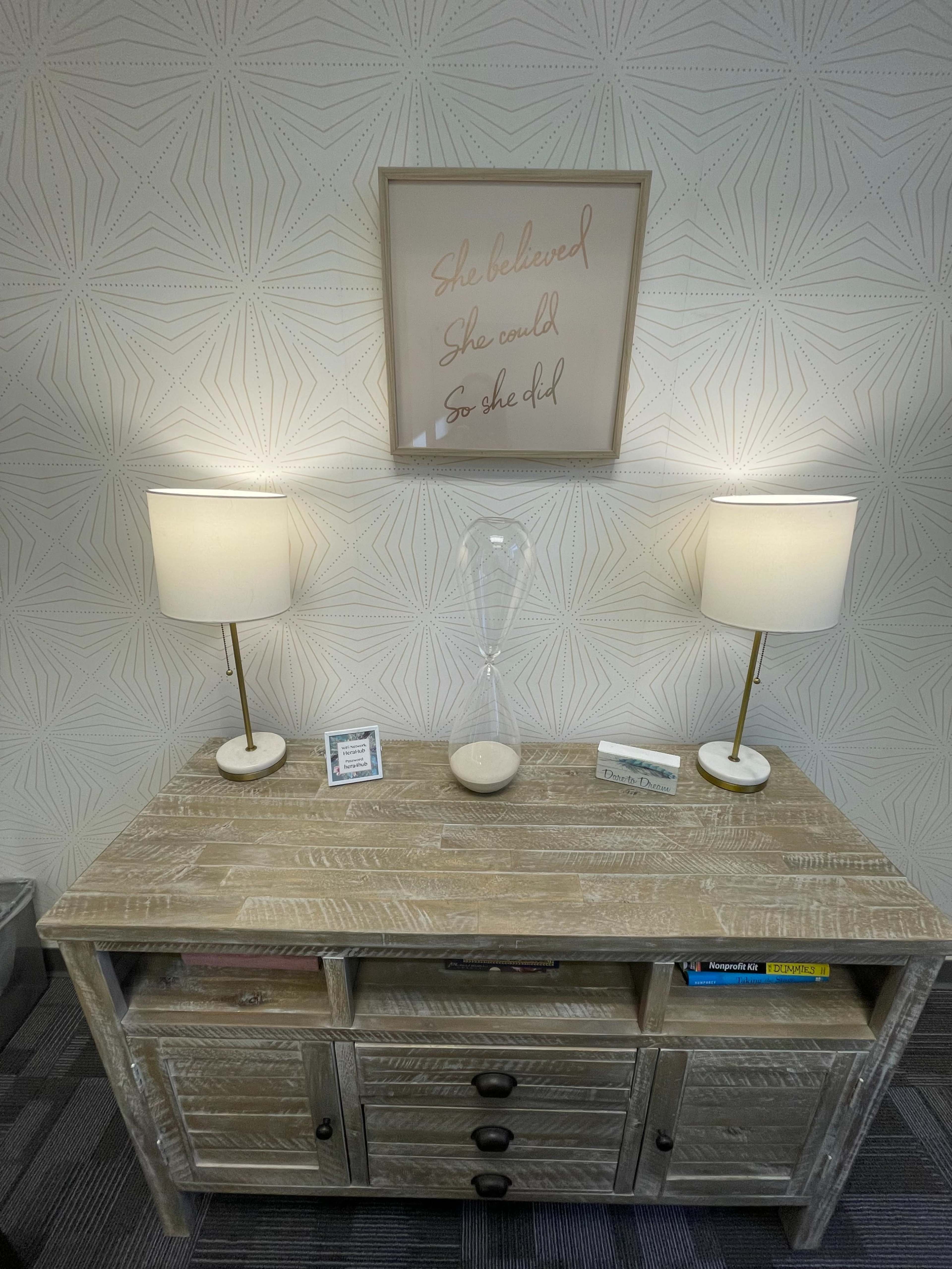A wooden console table with two lamps, an hourglass, and a framed inspirational quote on the wall above it.