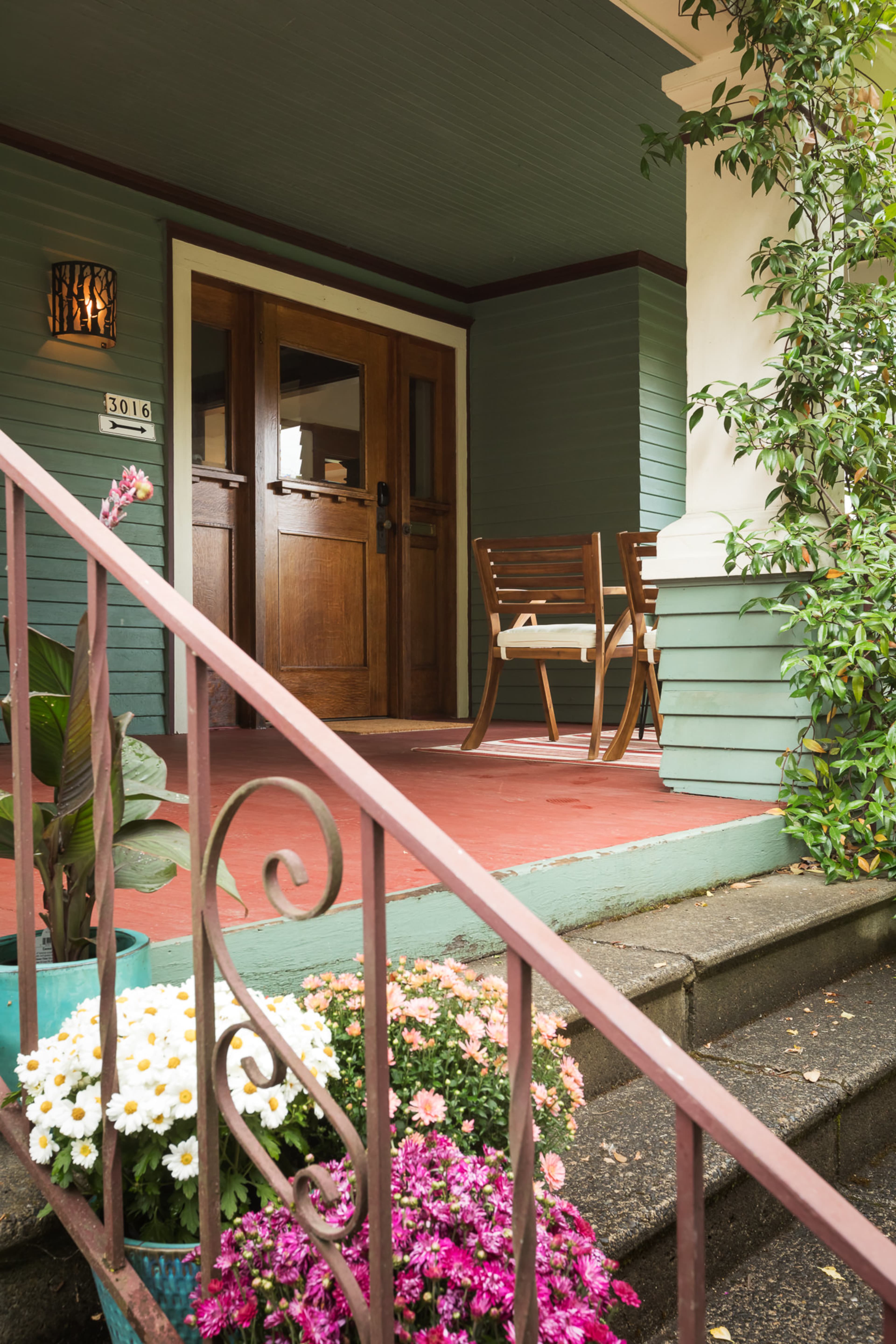 The entrance features a wooden door framed by green walls, with a small set of stairs leading to a porch adorned with potted flowers and seating.
