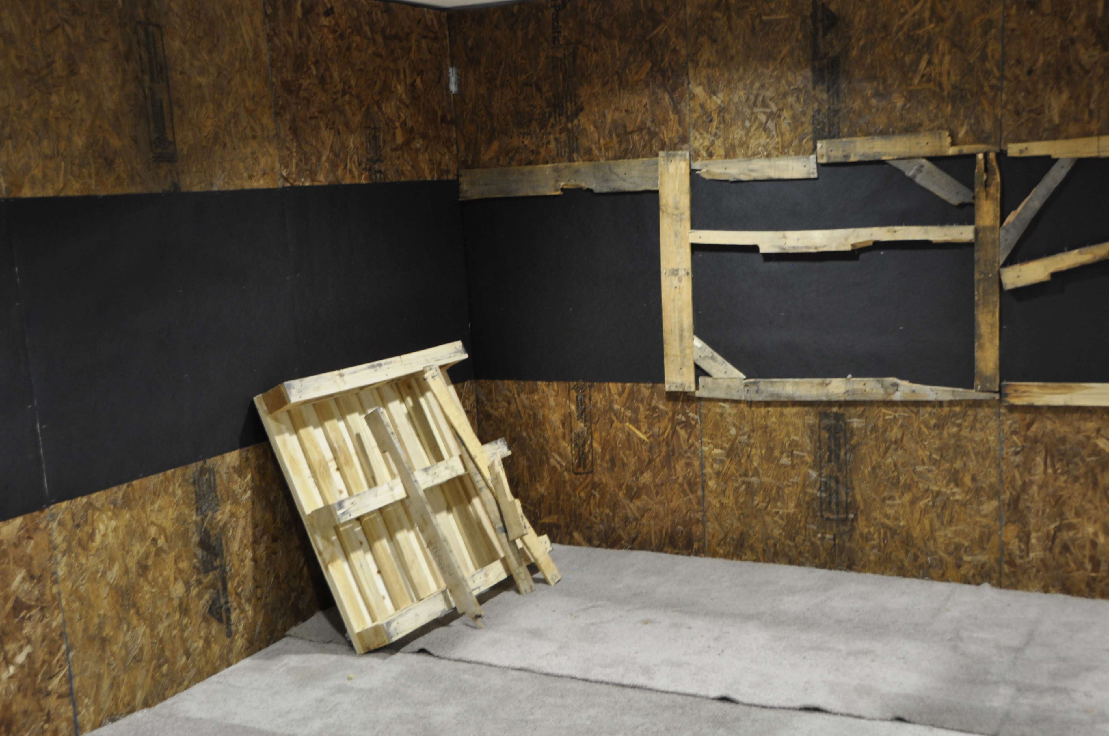 The image shows an interior room with wooden walls, a gray carpeted floor, and a pile of wooden pallets leaning against the wall.