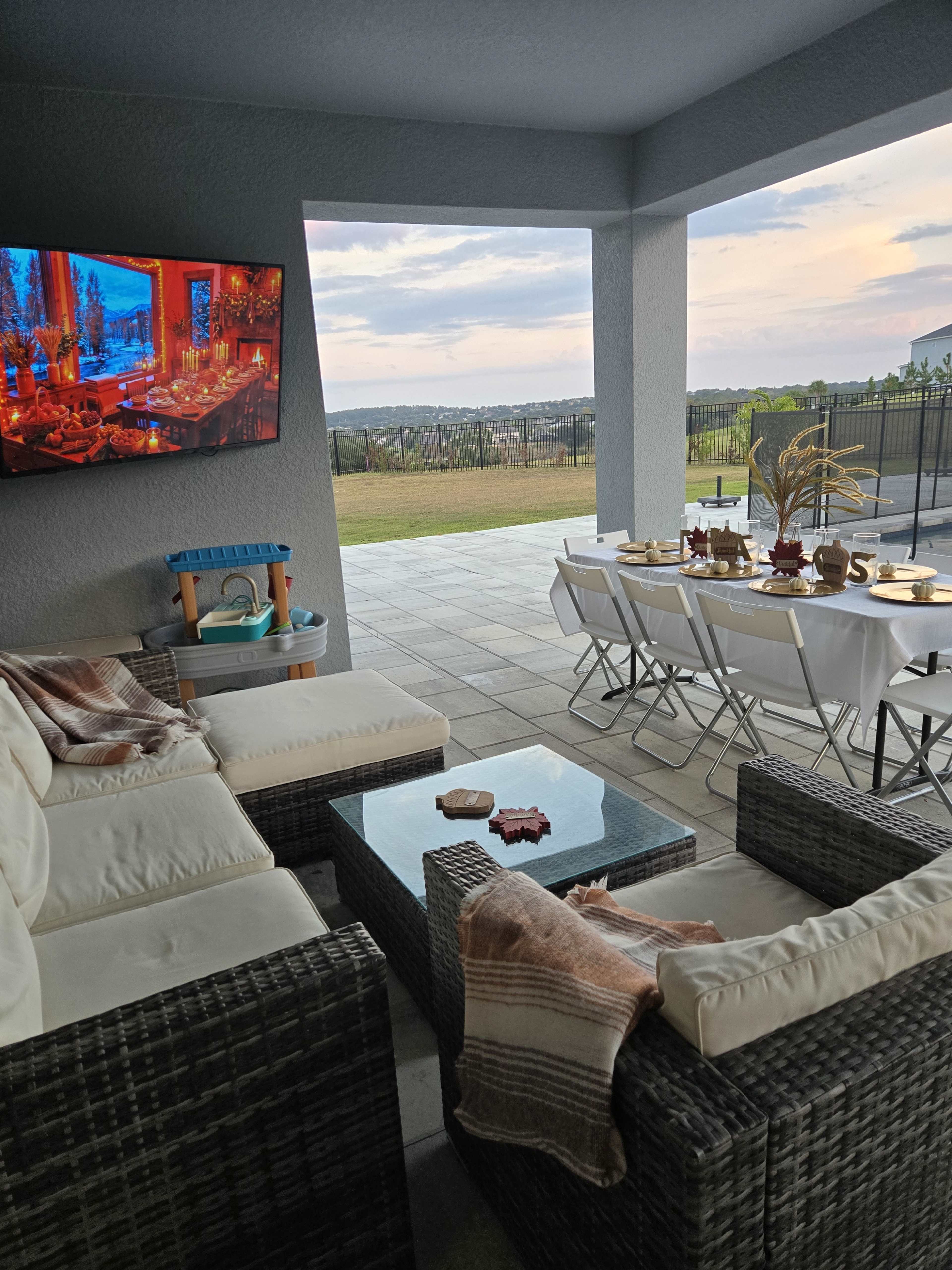 A cozy outdoor patio features a seating area with a television, a table set for dining, and a scenic view of the landscape beyond.