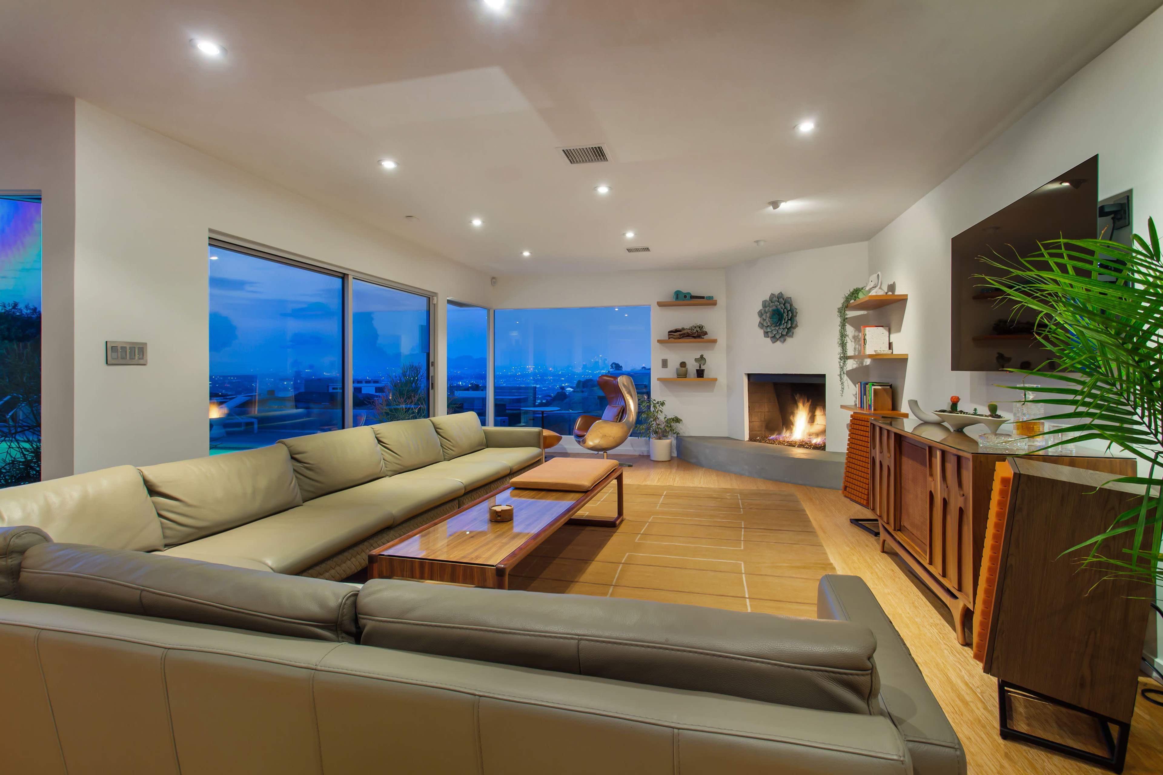 A modern living room features a large sectional sofa, a wooden coffee table, and a fireplace, with large windows offering a view of the skyline.