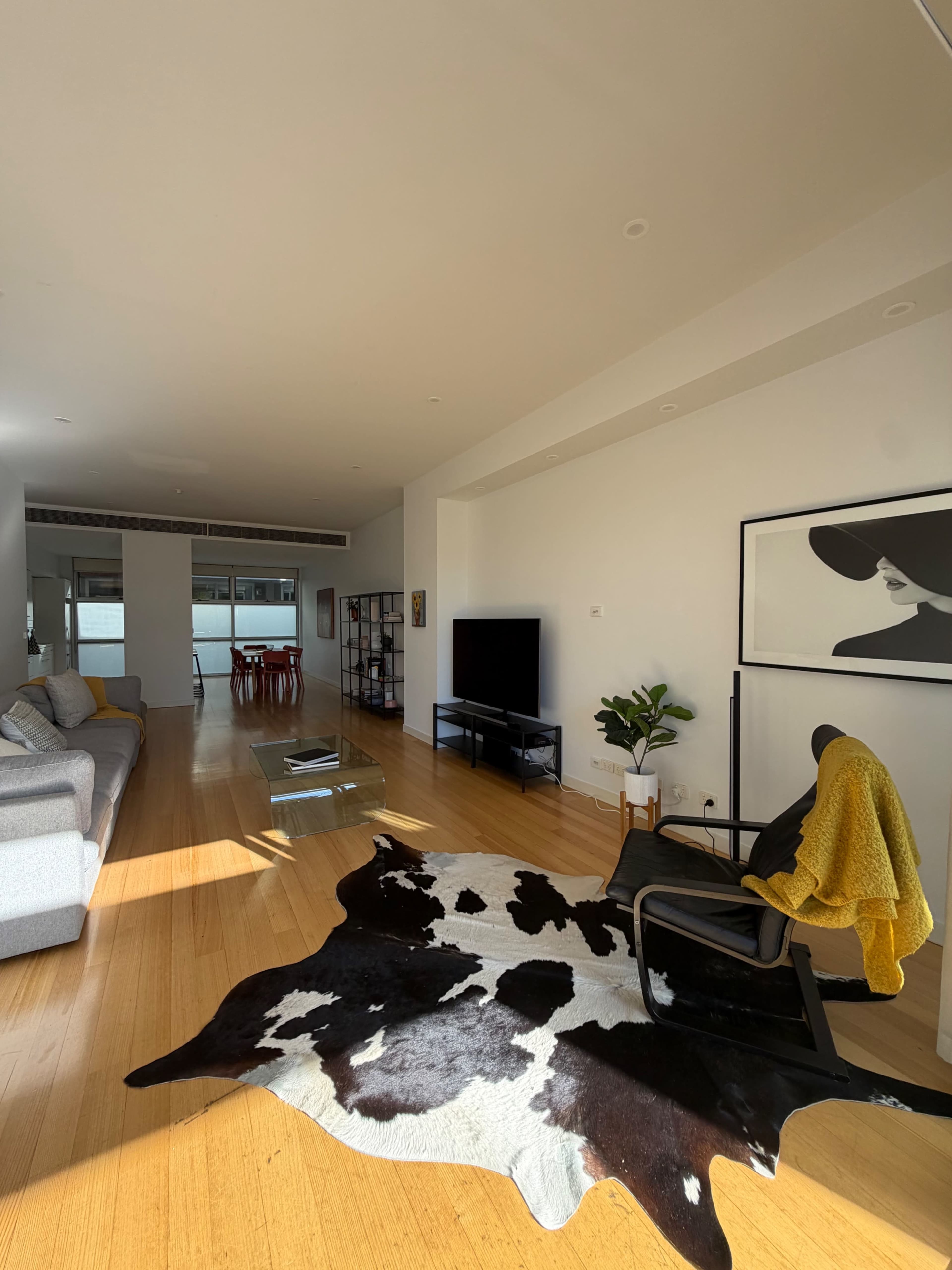 The image shows a spacious living room featuring a gray sofa, a black chair with a yellow throw, a cowhide rug, and a television against a white wall, with a dining area visible in the background.