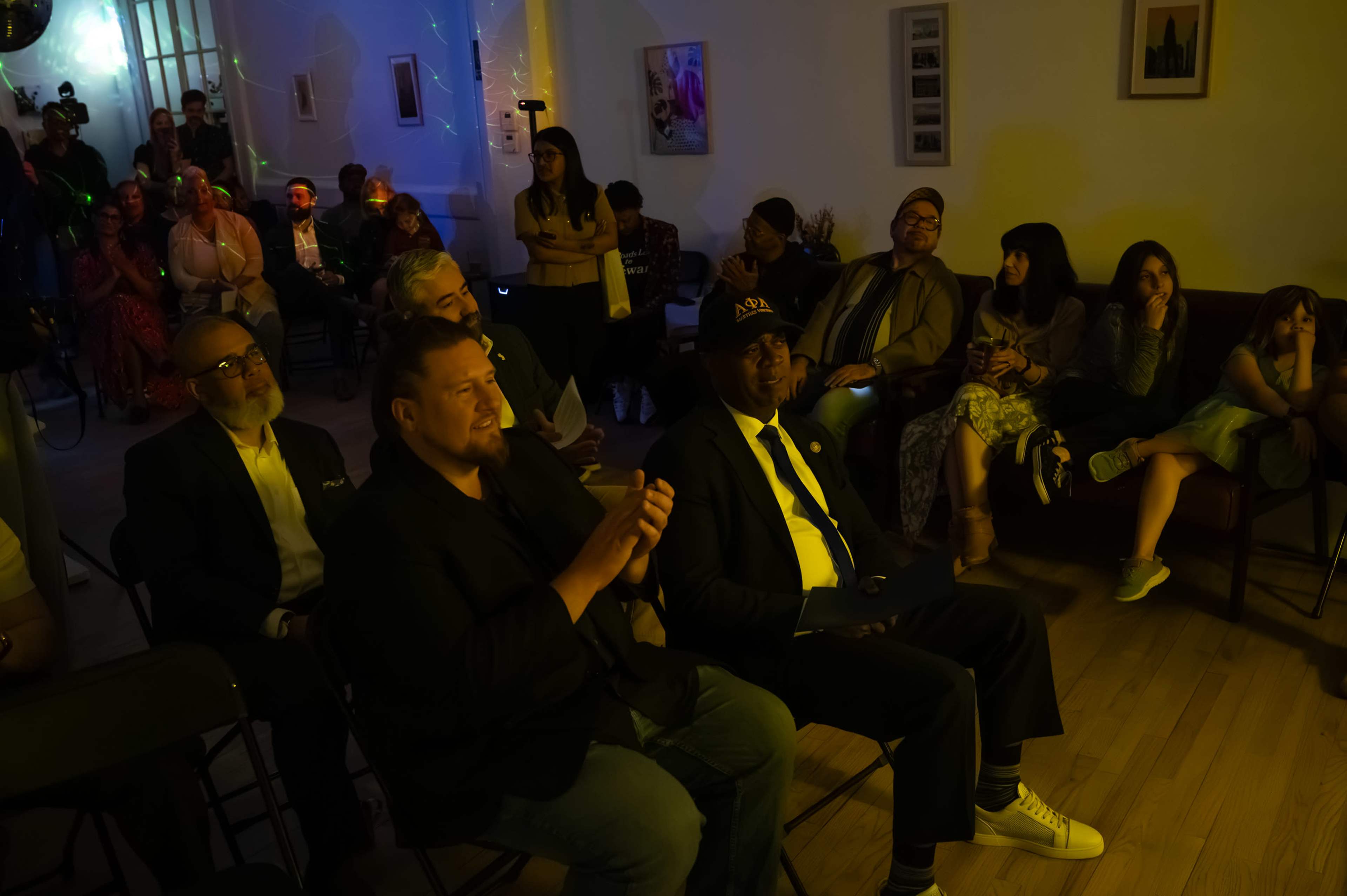 A diverse group of people sits attentively in folding chairs, watching an event in a softly lit room with artwork on the walls.