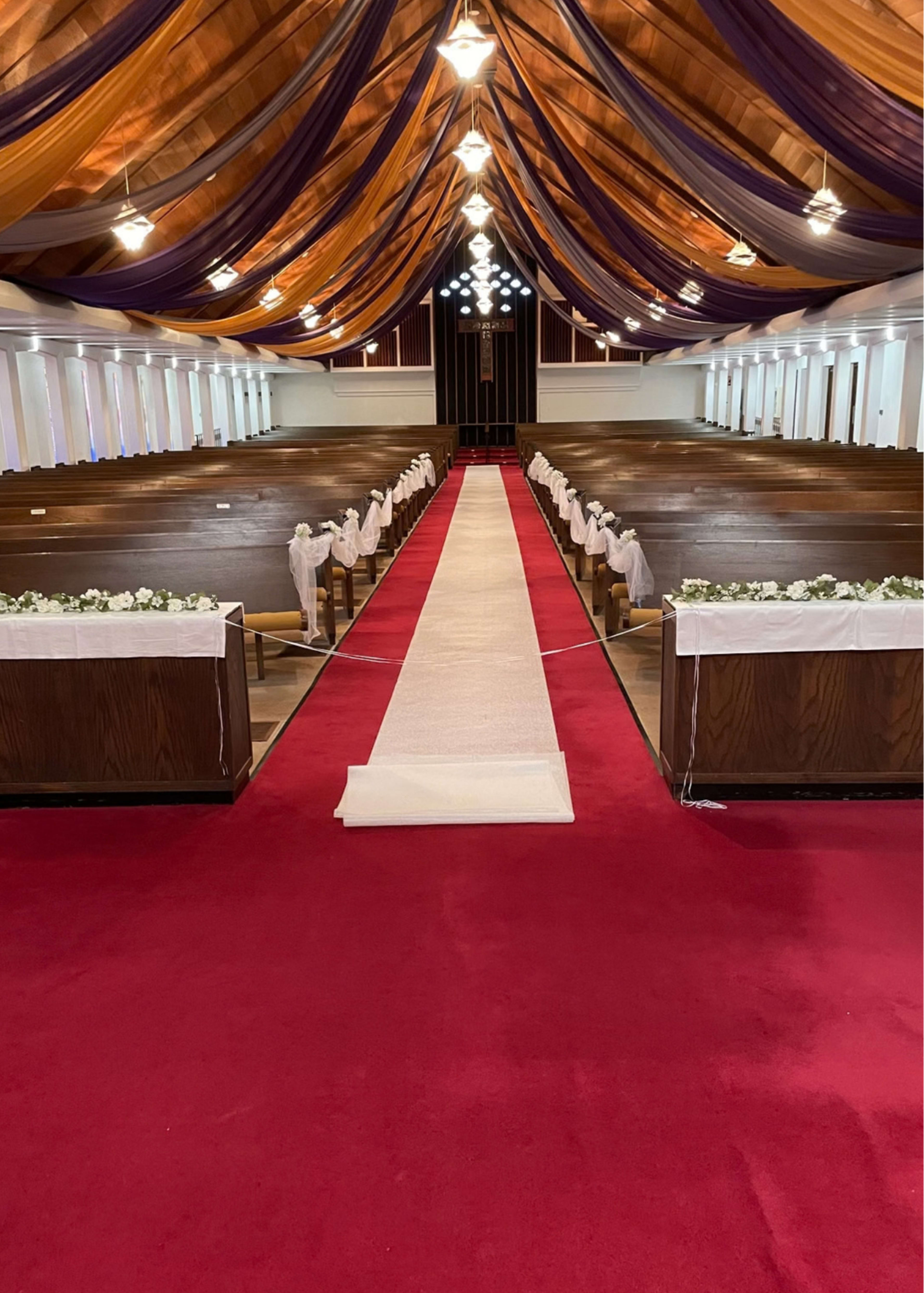 The image shows a decorated interior of a church with wooden pews along the sides, a red carpet down the aisle, and a floral arrangement at the front.