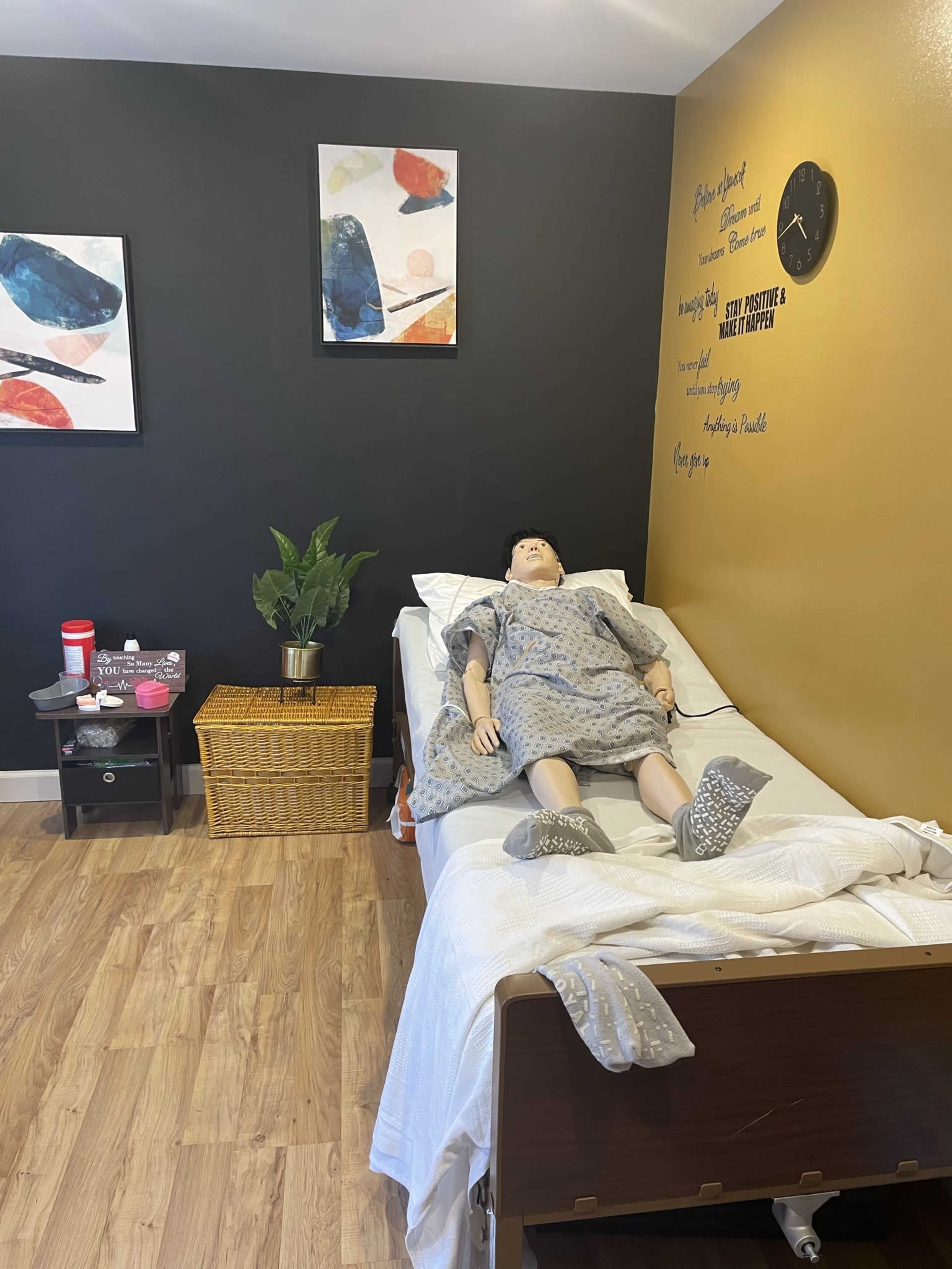 A simulation room with a mannequin lying in a hospital bed, surrounded by a small table, a basket, a plant, and framed artwork on the wall.