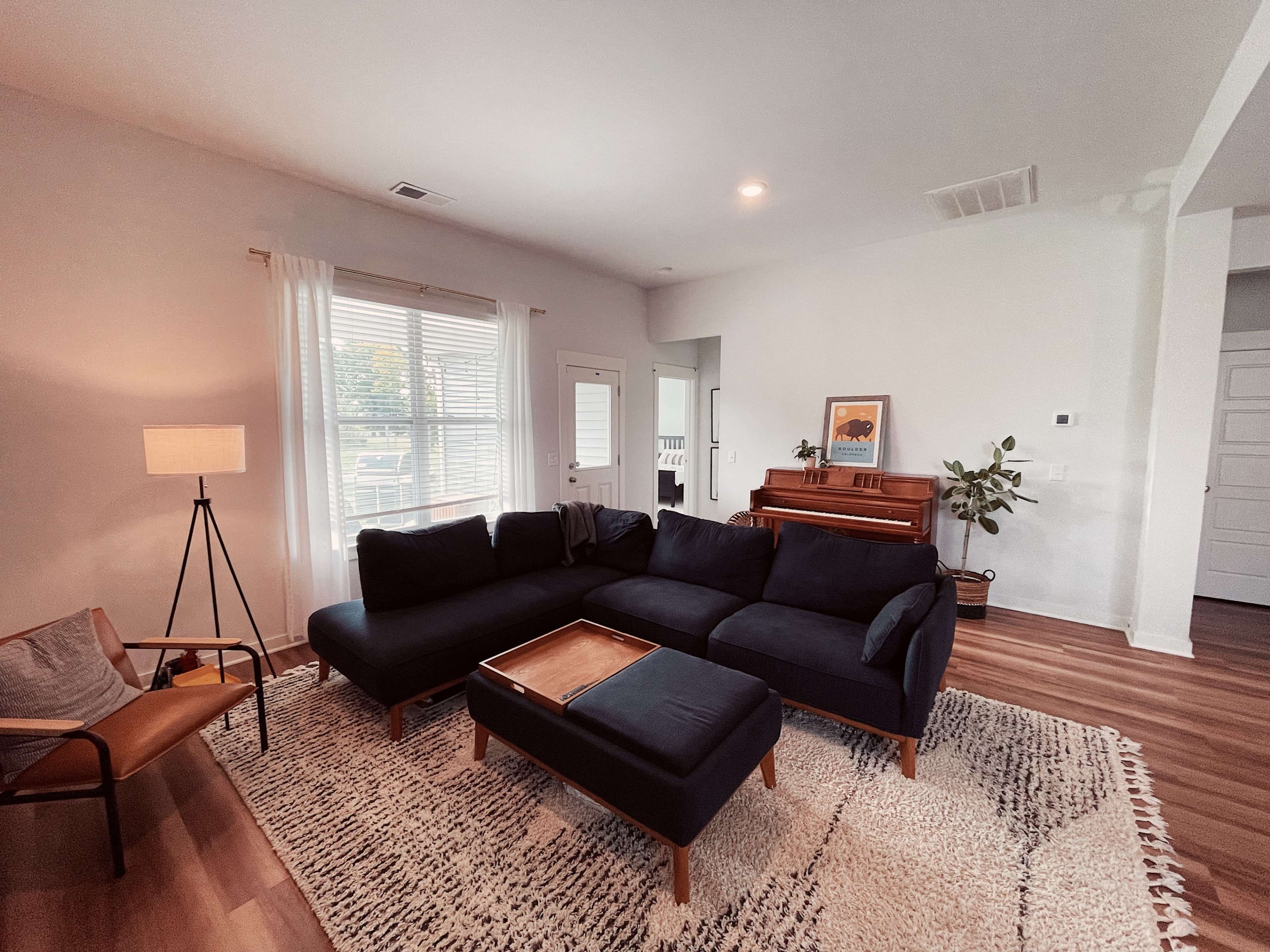A living room features a dark sectional sofa, a wooden coffee table, a lamp, and a piano, with windows allowing natural light to enter.