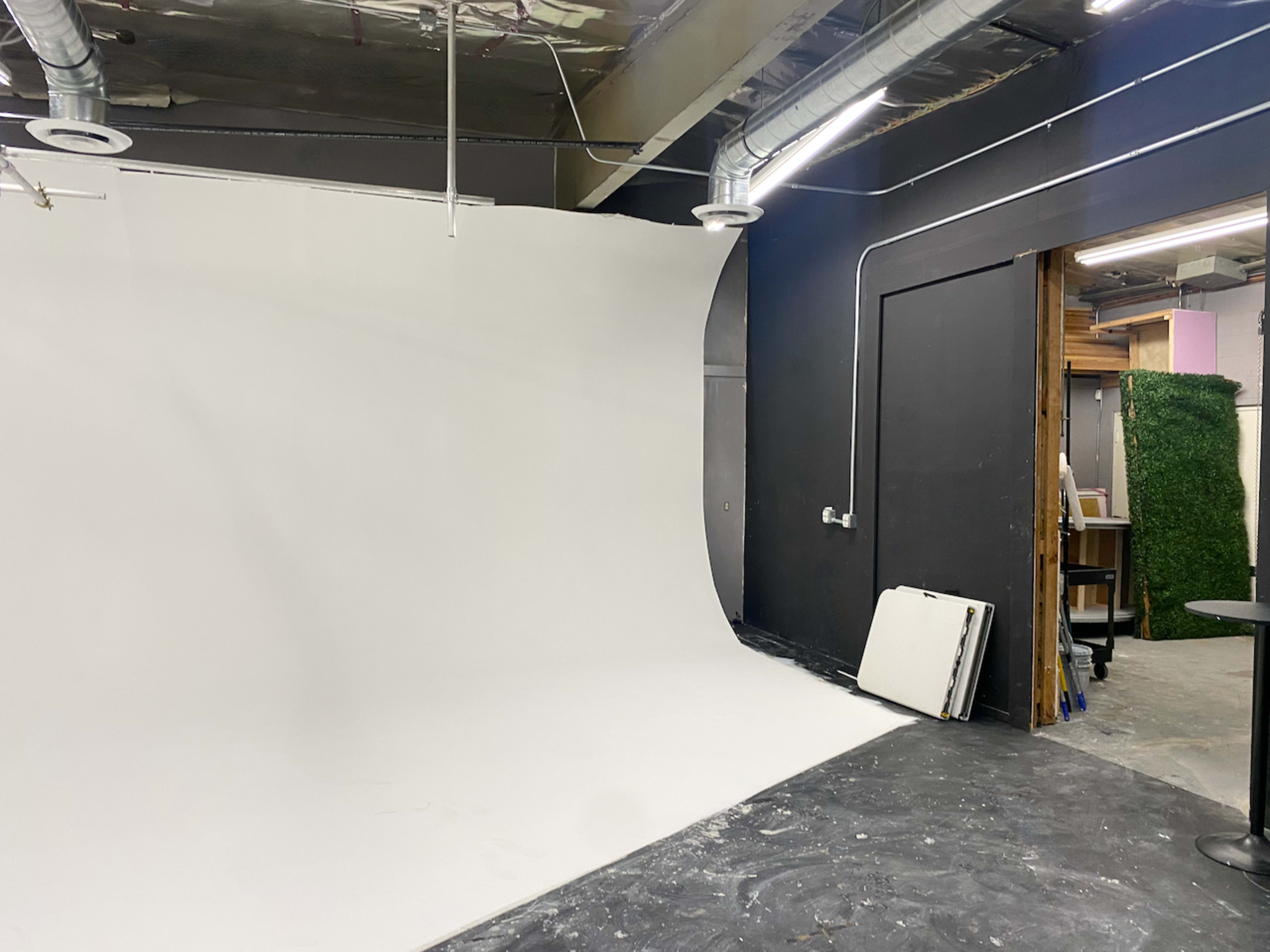 The image shows a spacious photography studio with a seamless white backdrop and a black wall, featuring metal ductwork and various equipment on the floor.