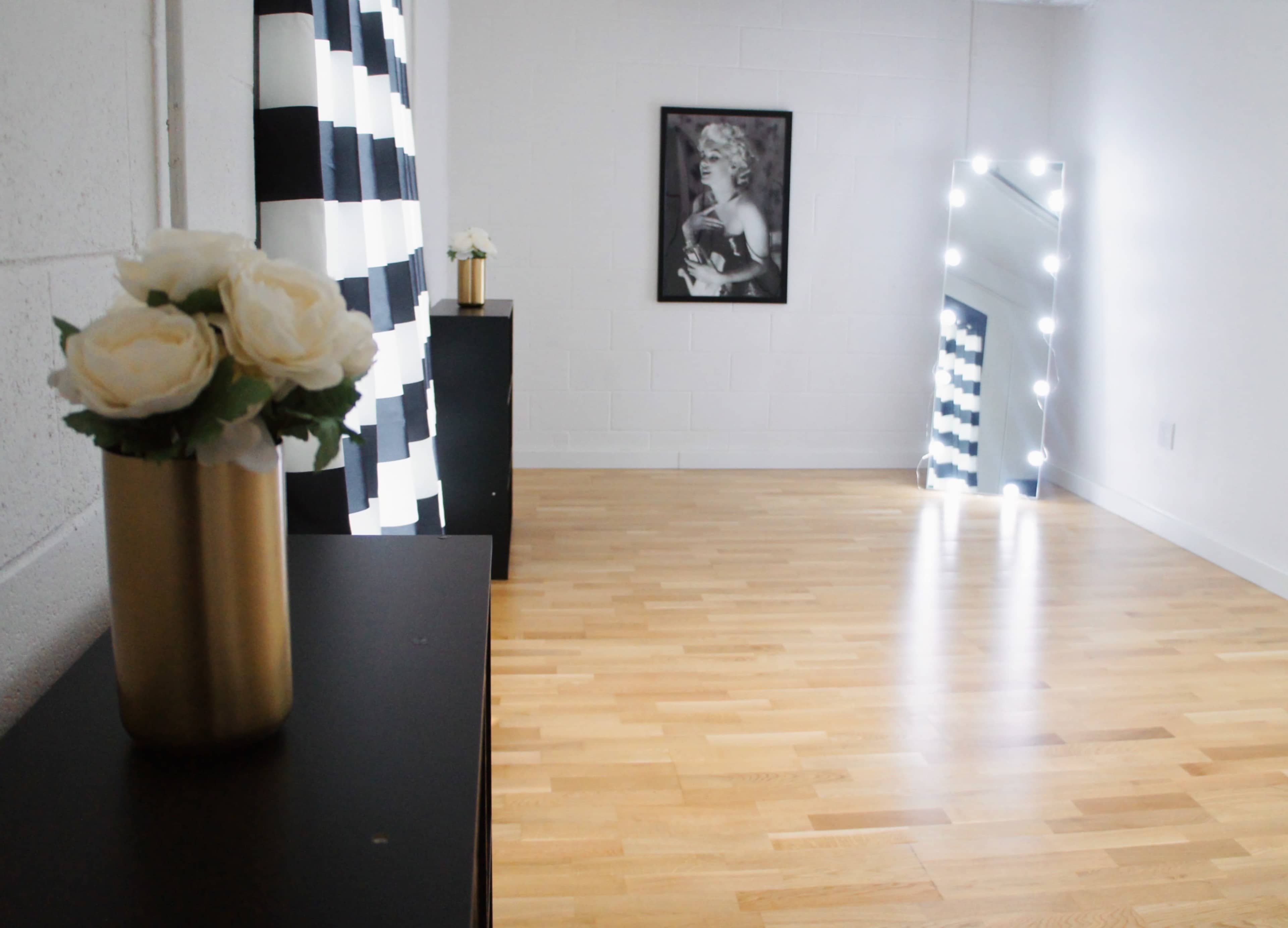 The image shows a brightly lit room with wooden flooring, a black dresser, a vase of roses, a large mirror with lights, and a black and white striped curtain on one wall.