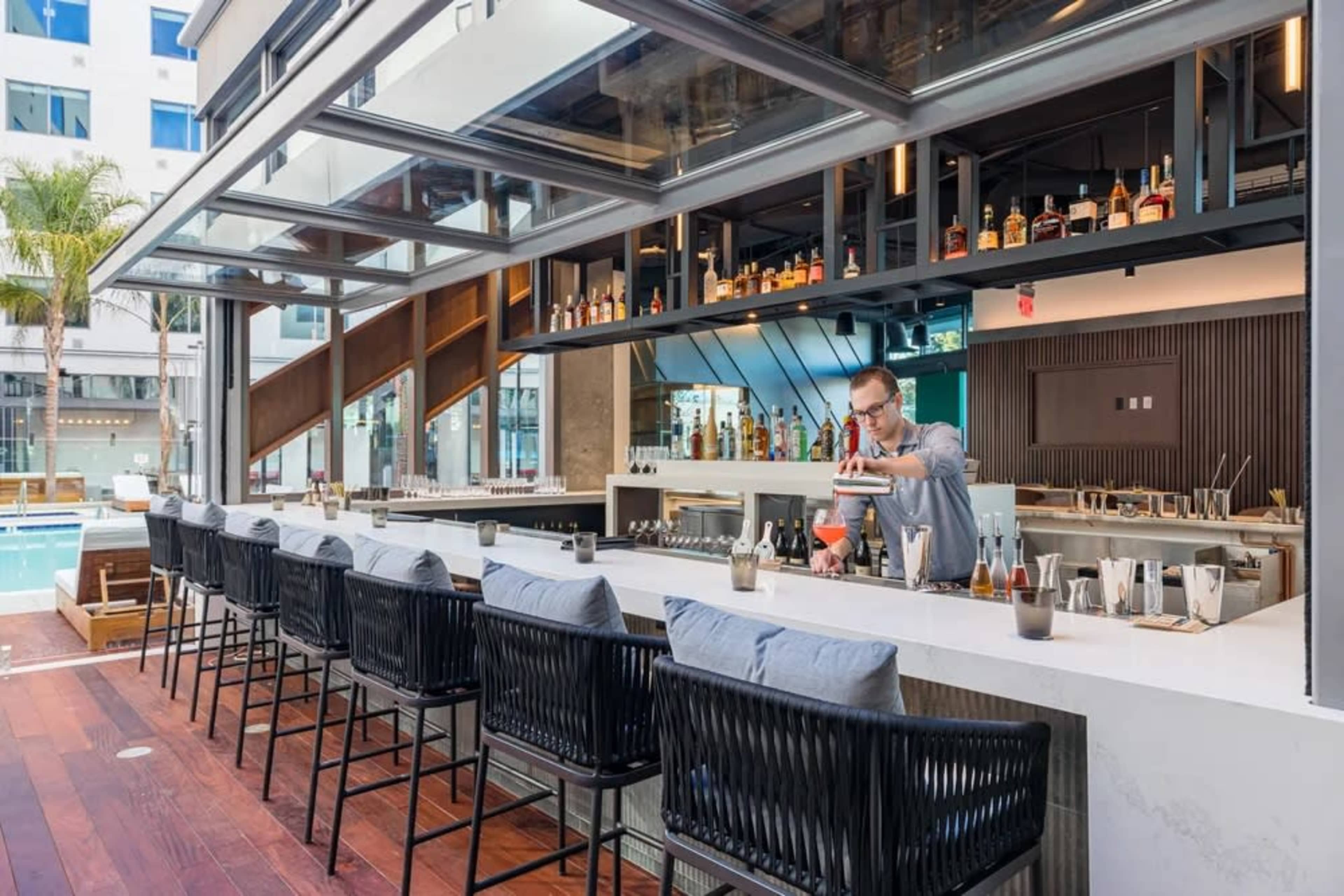 A bartender prepares drinks at a modern outdoor bar that overlooks a swimming pool.