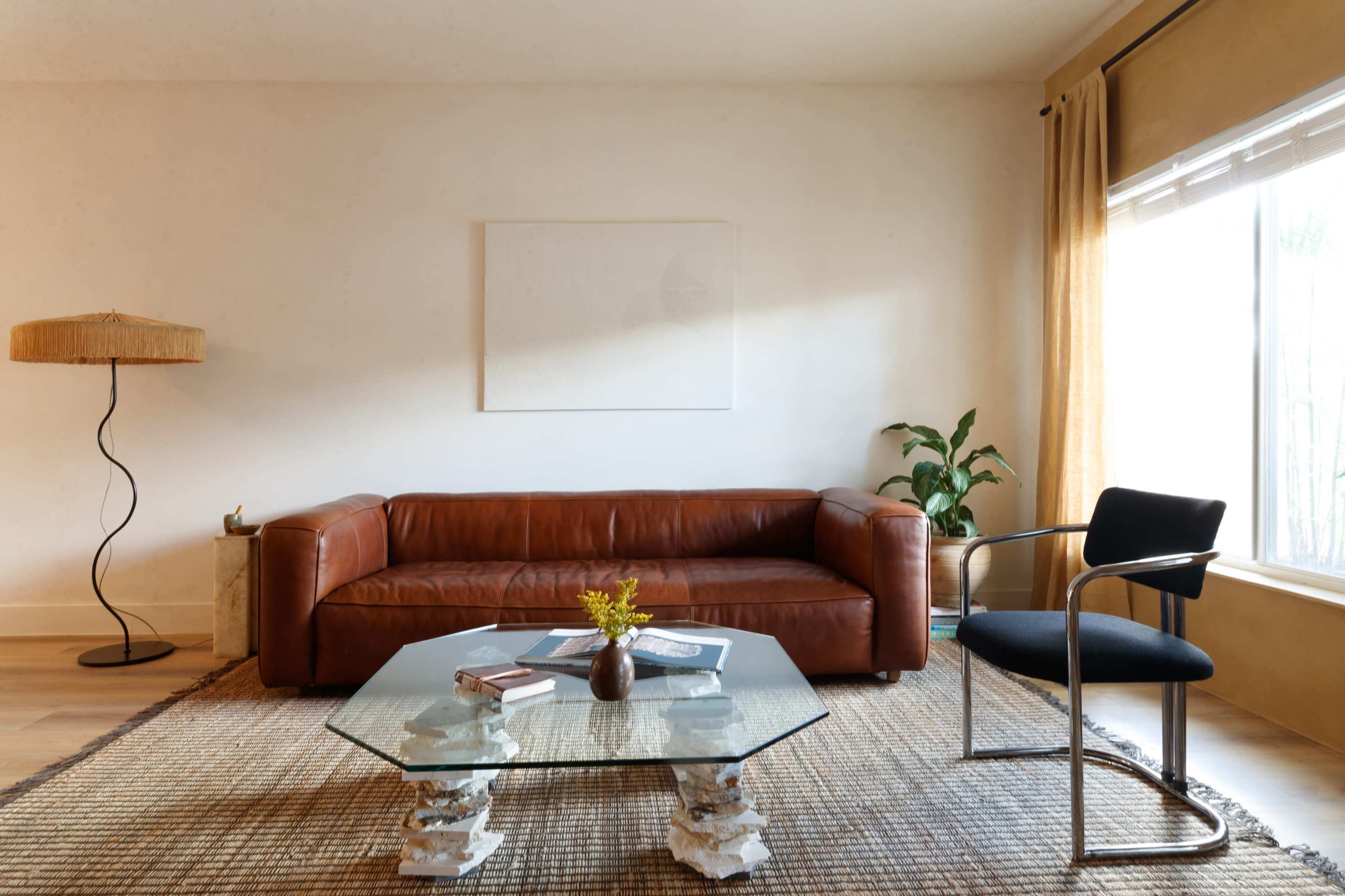 The image shows a modern living room with a brown leather couch, a glass coffee table, a black chair, and a floor lamp beside a large window with sheer curtains.