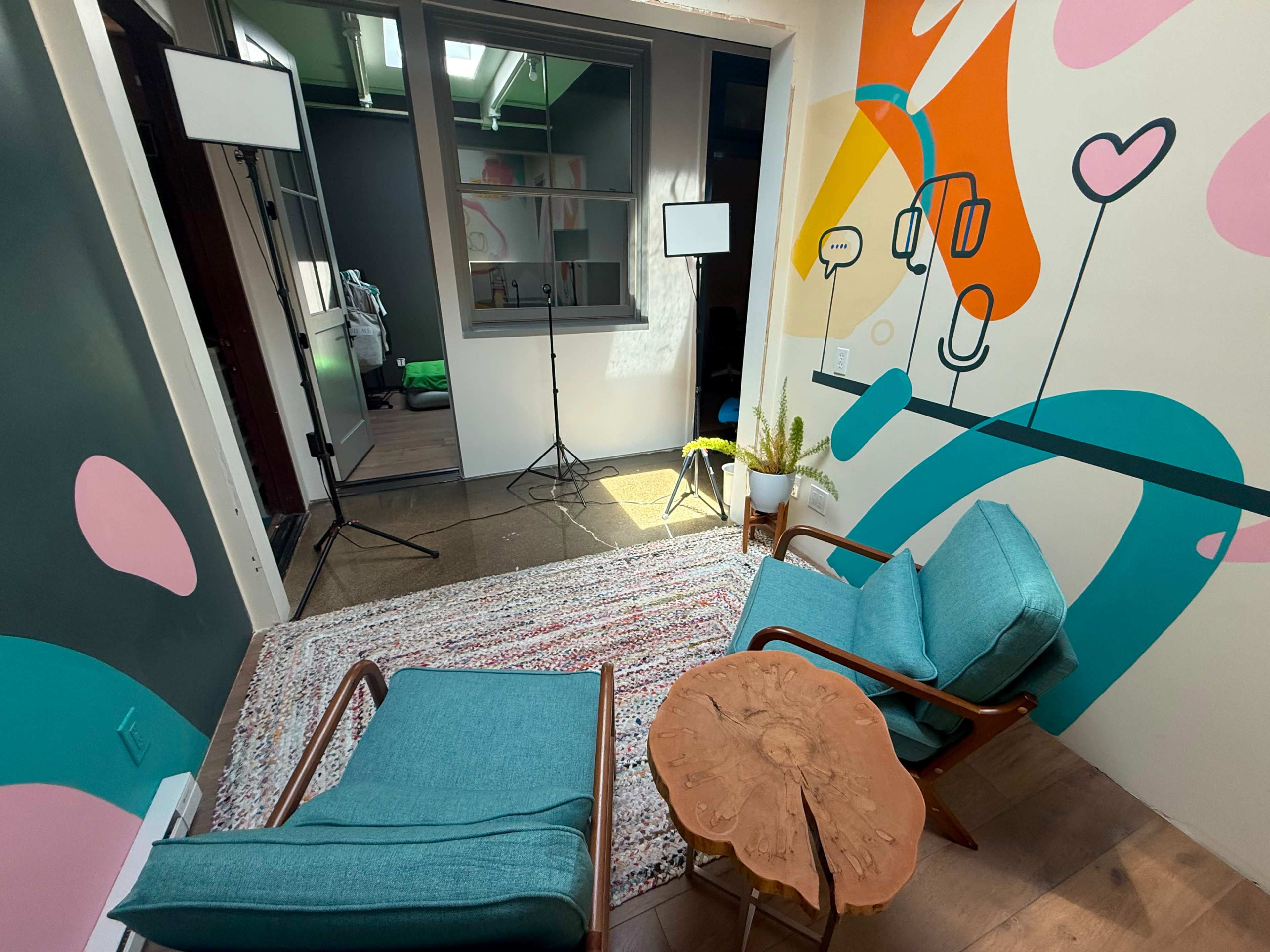 The image shows a small, brightly decorated room featuring two turquoise chairs, a wooden coffee table, and colorful wall murals, with soft lighting equipment positioned nearby.