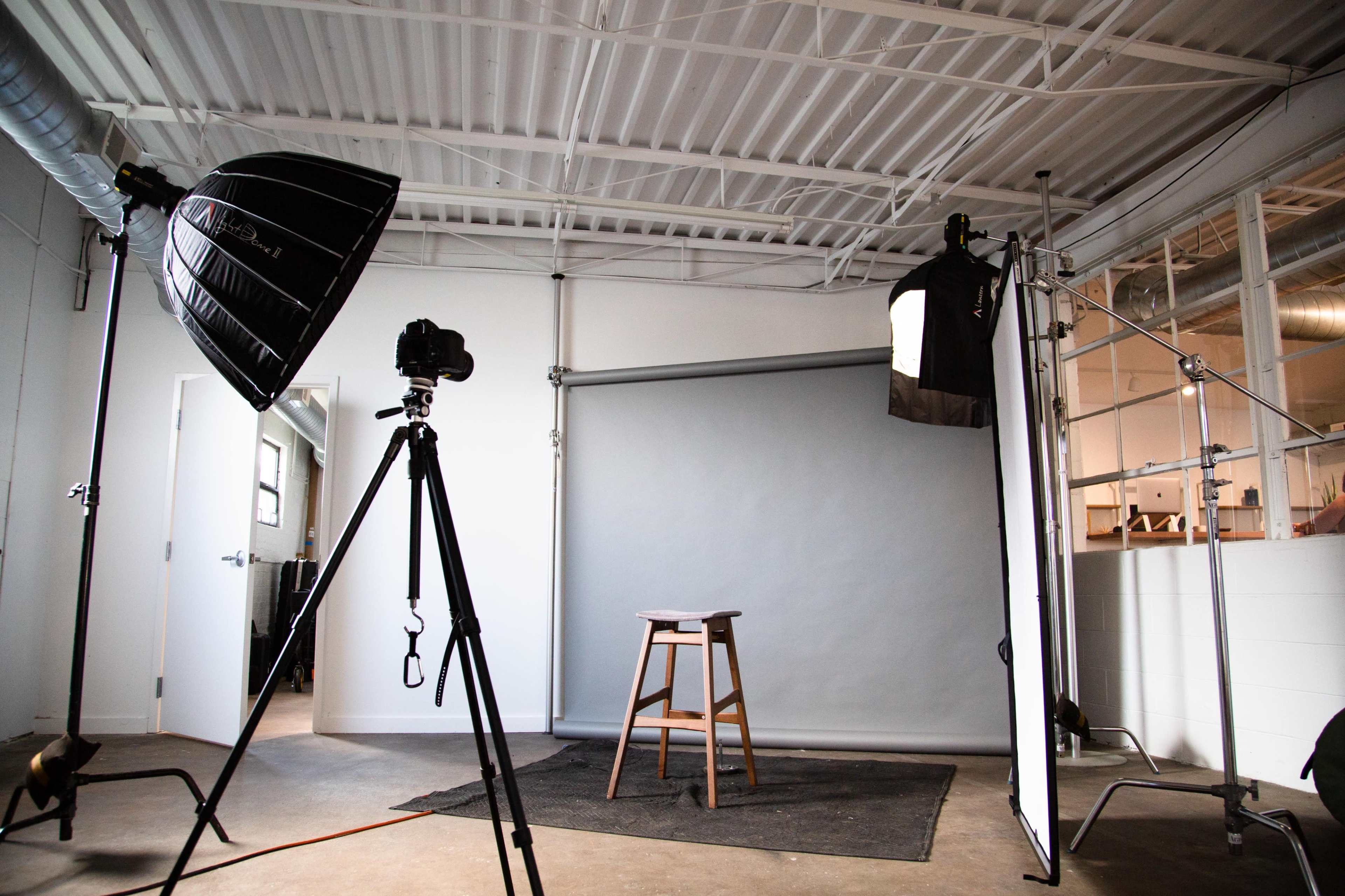 A photography studio with a camera on a tripod, a stool, and lighting equipment set up against a gray backdrop.