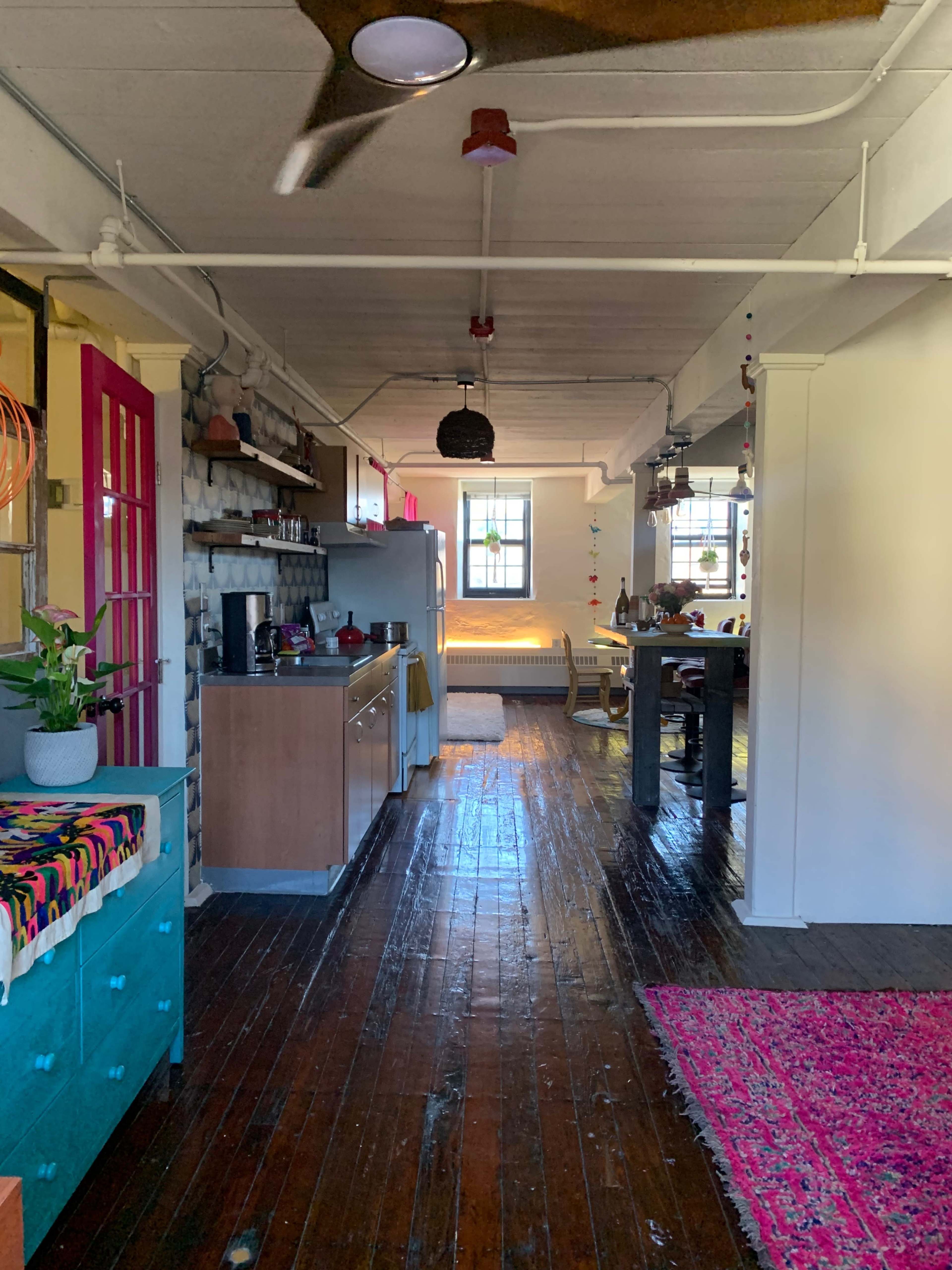 A long, narrow kitchen and dining area with wooden floors, featuring a mix of colorful cabinets, a stainless steel refrigerator, and a dining table set against a bright backdrop of windows.
