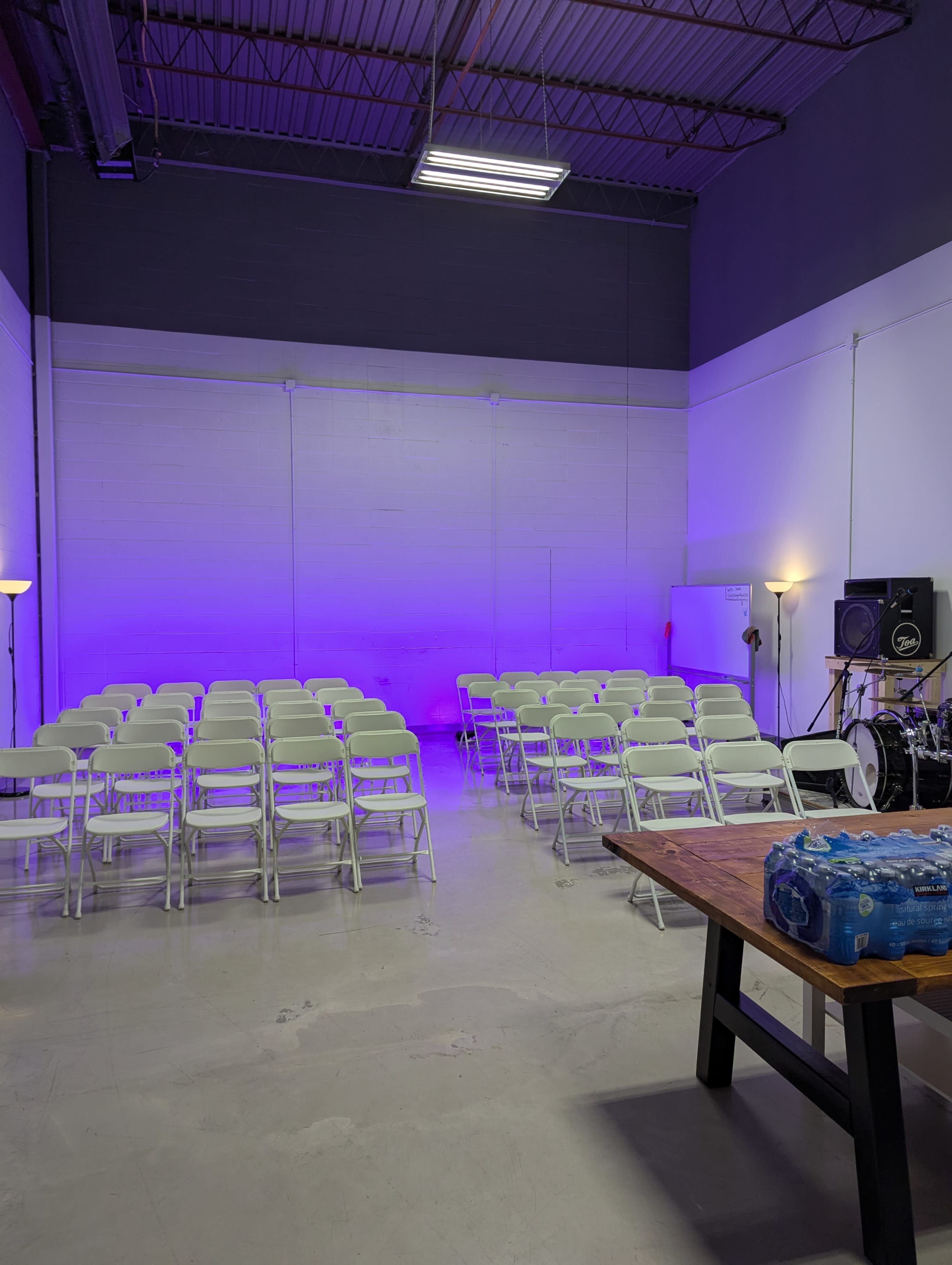 A room with white walls contains rows of folding chairs, a table with bottled water, and a music setup with speakers and instruments.