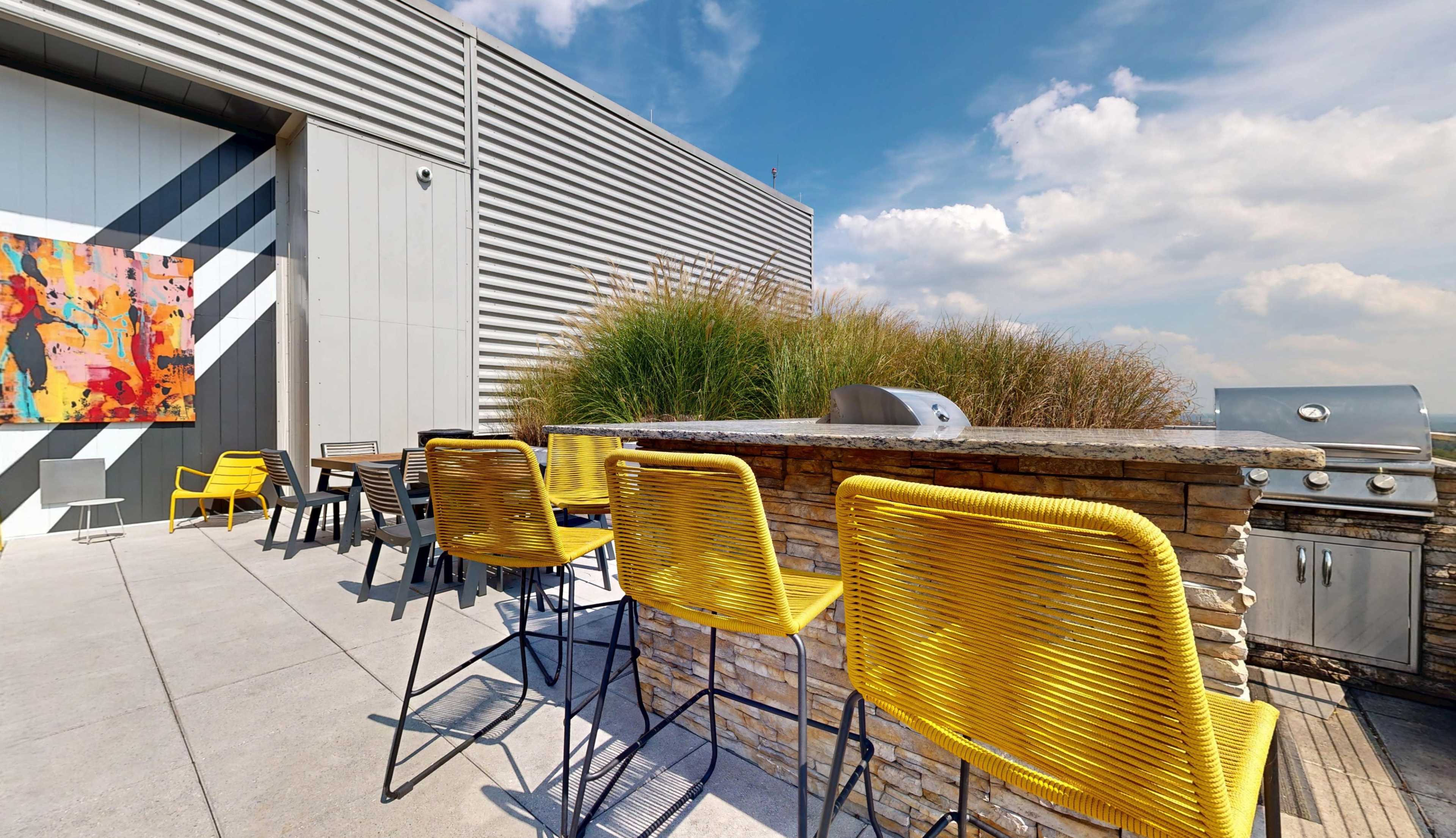 The image shows a modern outdoor seating area with yellow chairs and a stone bar, set against a backdrop of a building and blue sky with clouds.