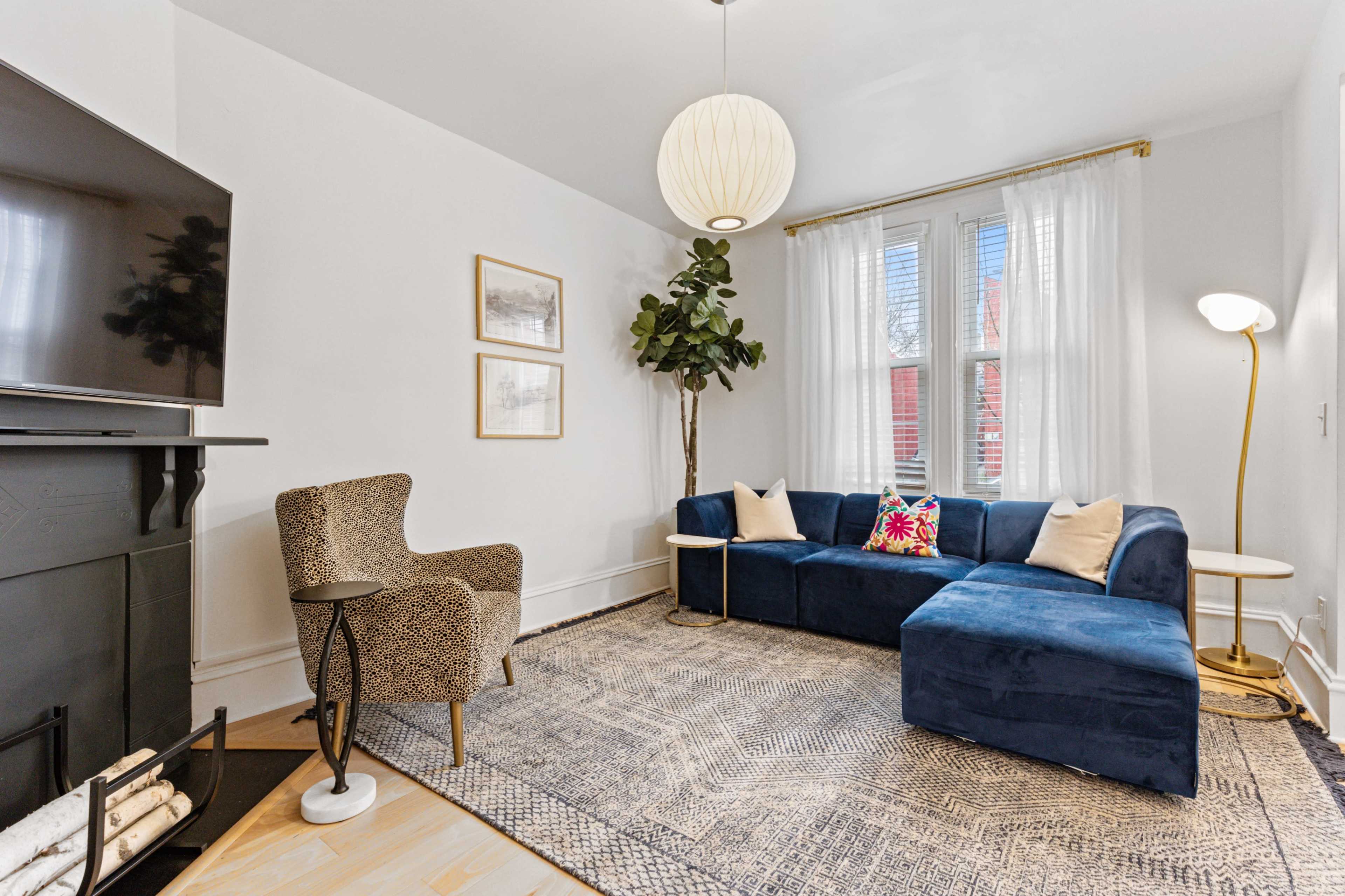 A modern living room features a blue sectional sofa, a patterned chair, a floor lamp, and a television mounted on the wall.