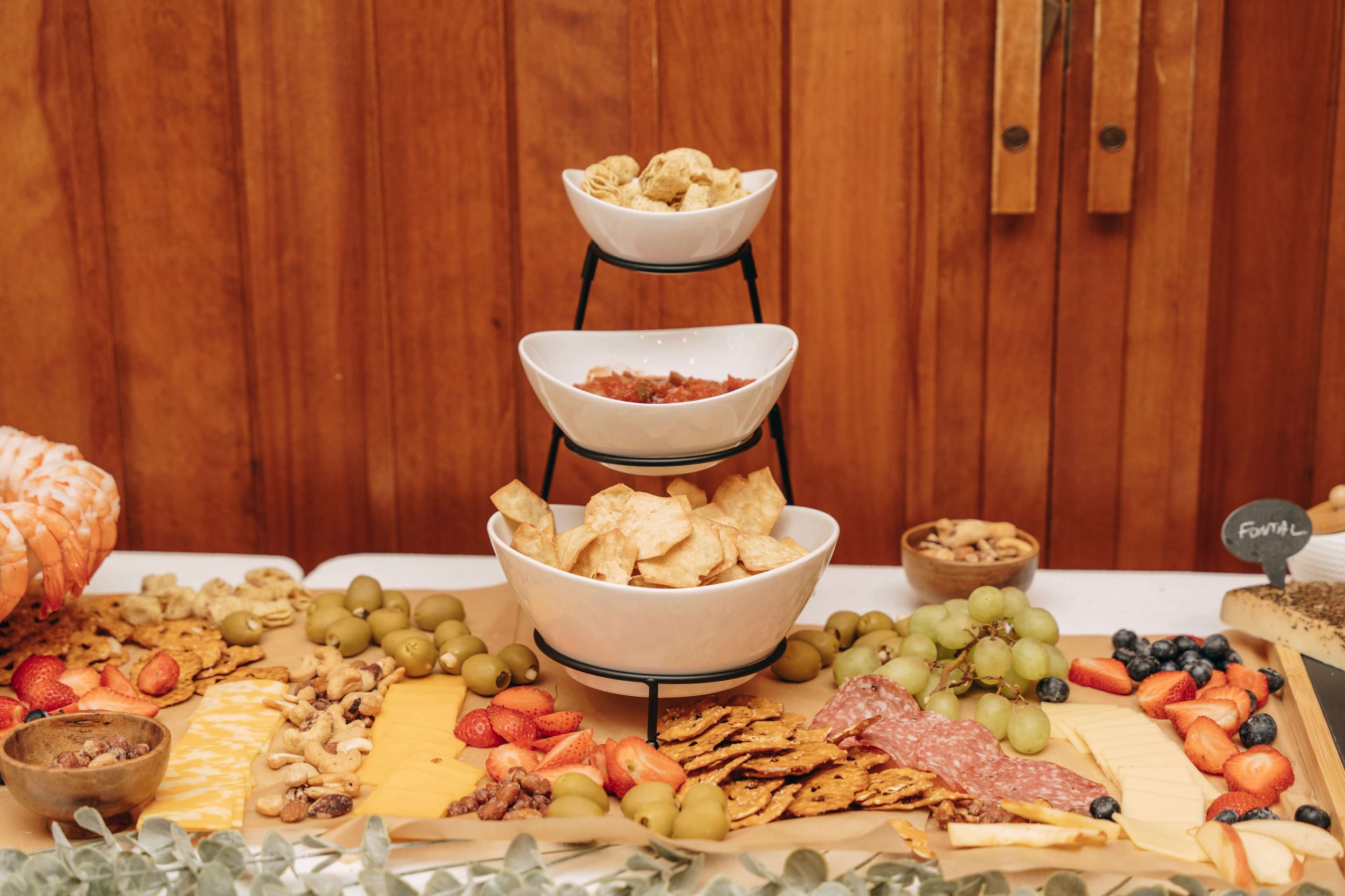 A tiered serving stand displays bowls of snacks, including chips and salsa, surrounded by an assortment of fruits, nuts, and crackers on a table.