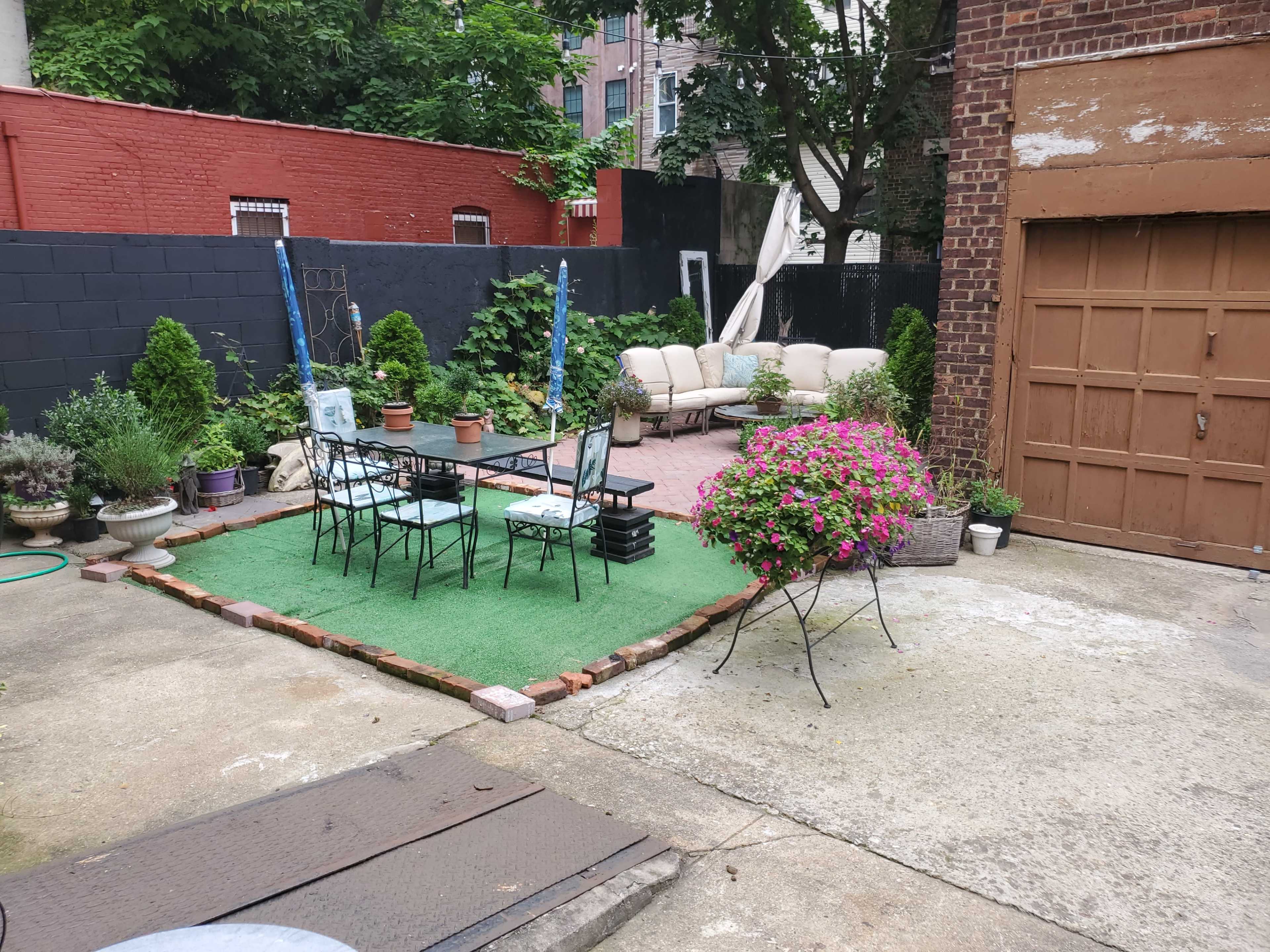 A backyard patio with a table and chairs on grass-like turf, surrounded by planters and a garage.