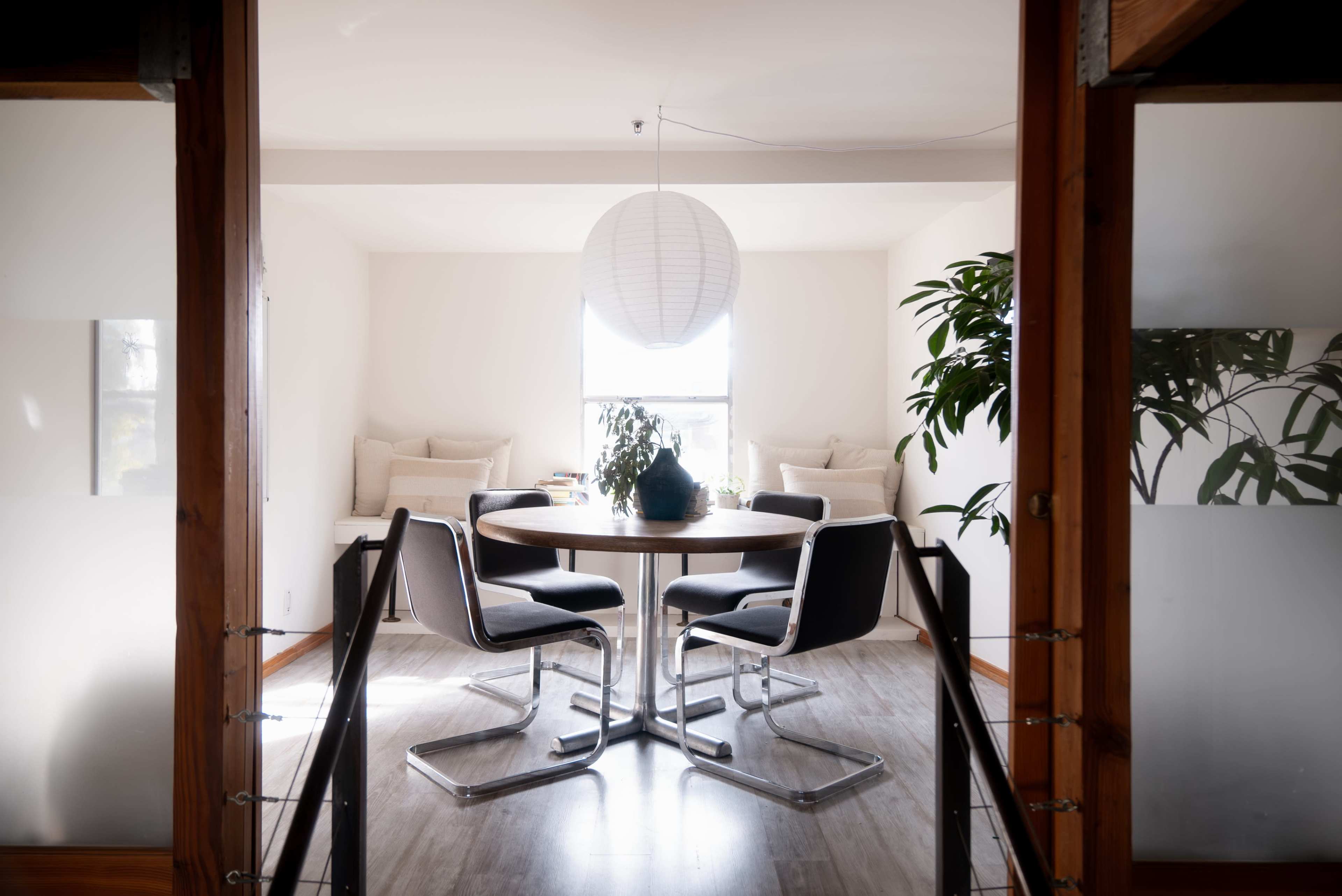 A round table with four chairs is set in a bright room, featuring a lantern overhead and plants in the corner.