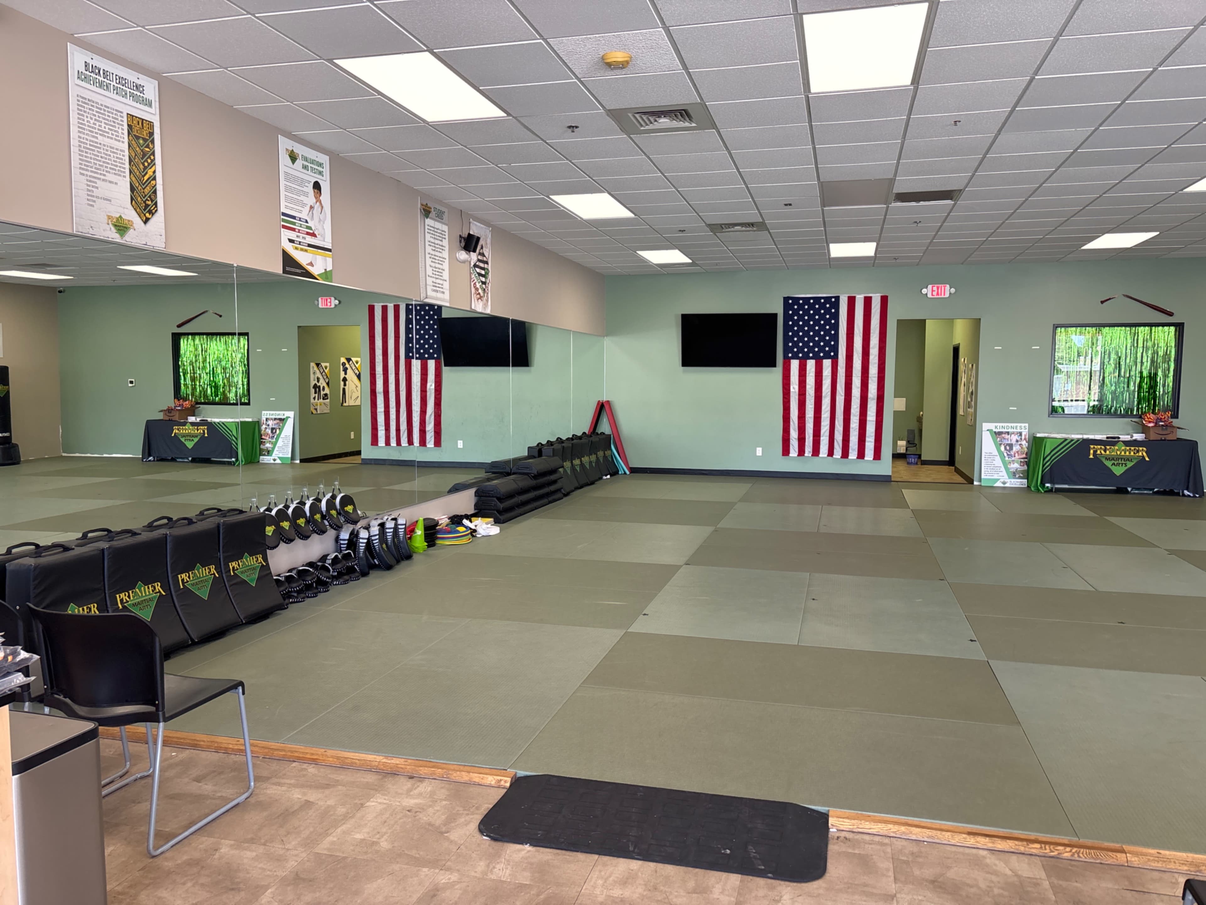 The image shows a martial arts training studio with green mats, two American flags on the wall, and equipment neatly arranged along one side.