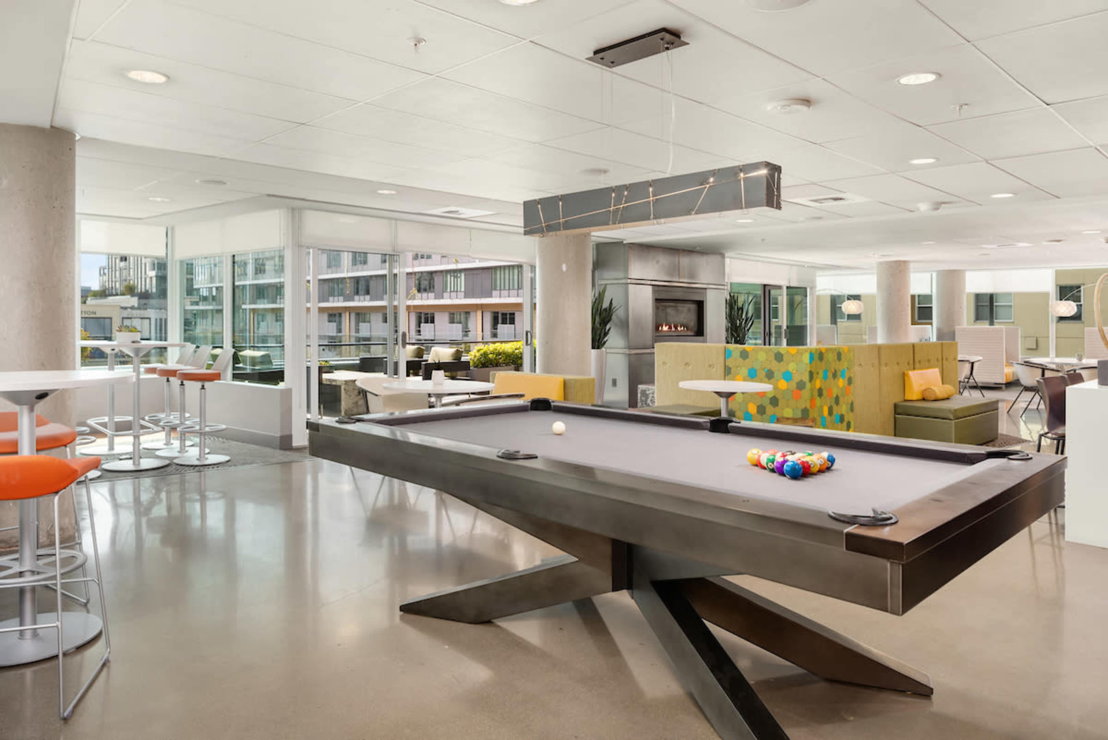 The image shows a modern lounge area featuring a sleek pool table, communal seating, large windows, and contemporary decor.