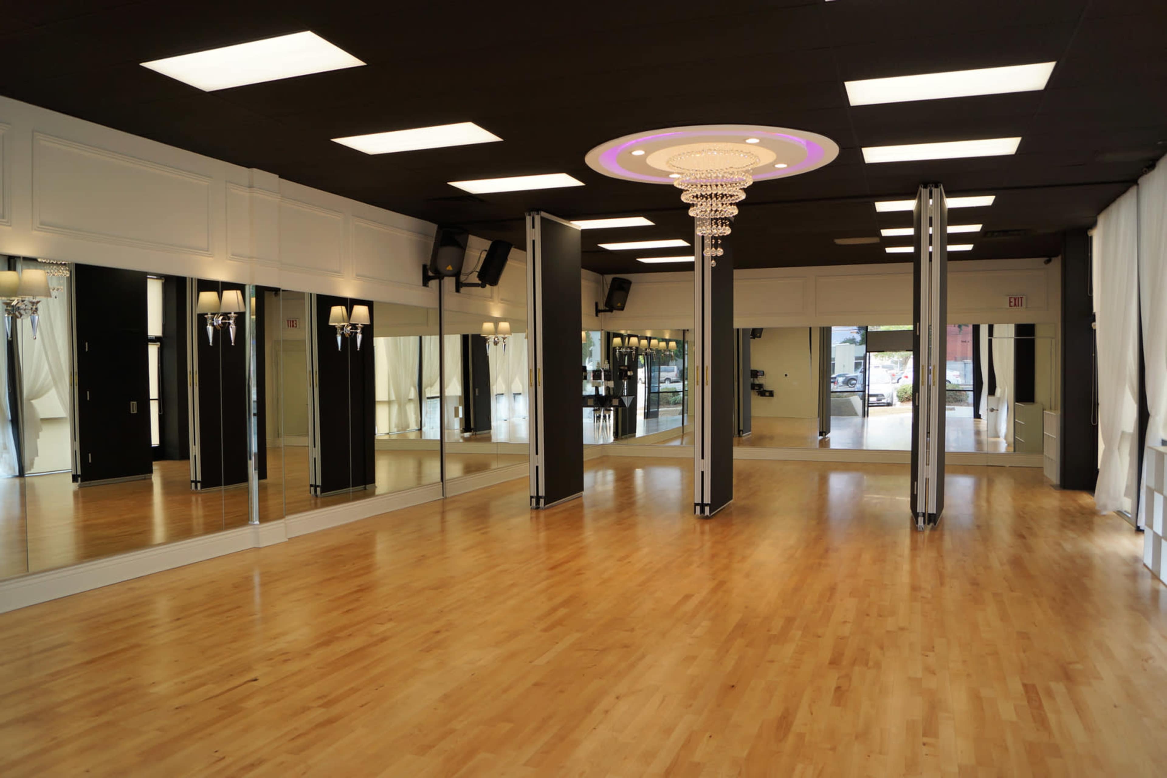 A spacious dance studio with hardwood floors, large mirrors, and a chandelier suspended from the ceiling.
