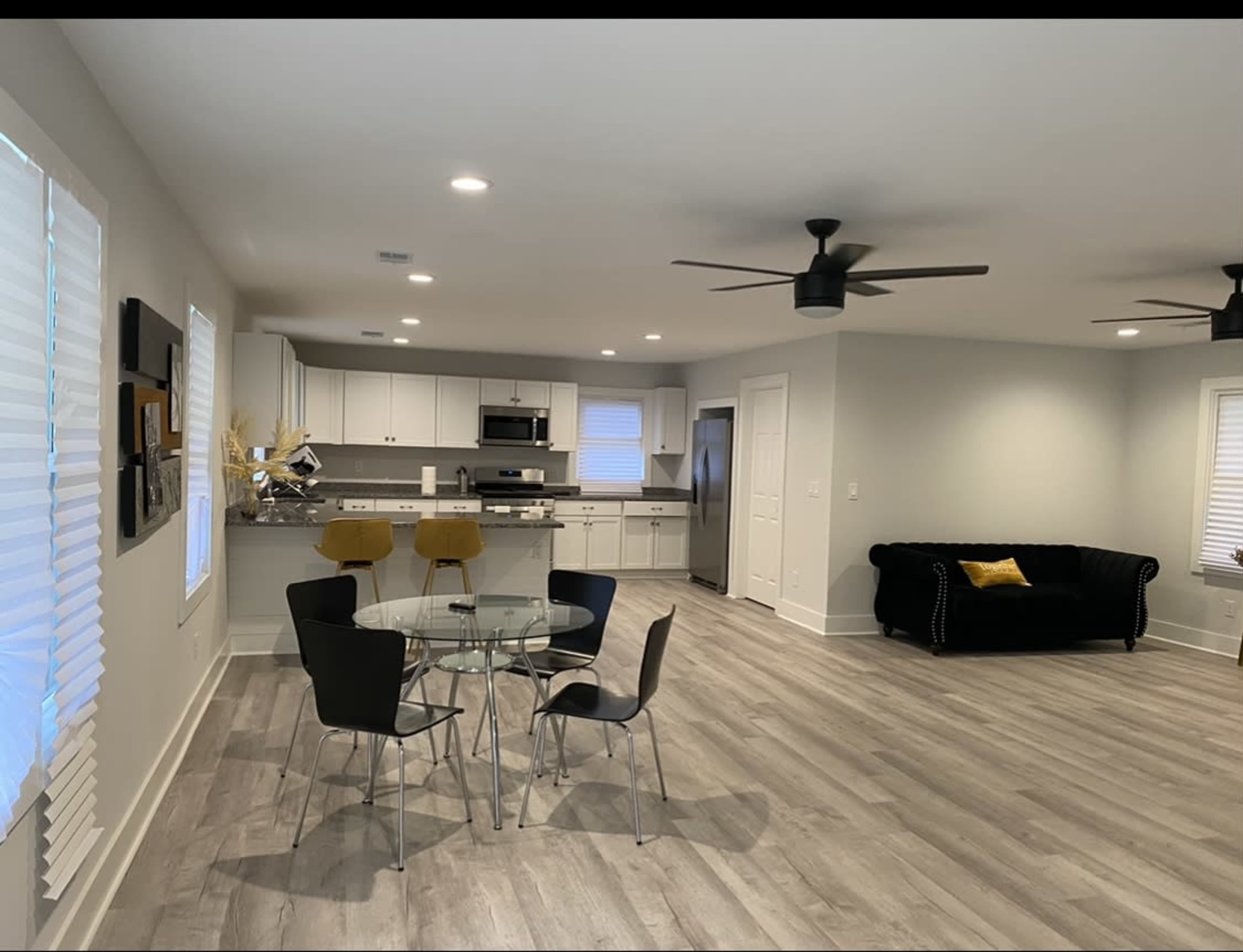 A modern, open-concept living space with a dining area featuring a glass table and four chairs, a kitchen with white cabinetry, and a living area with a black sofa.