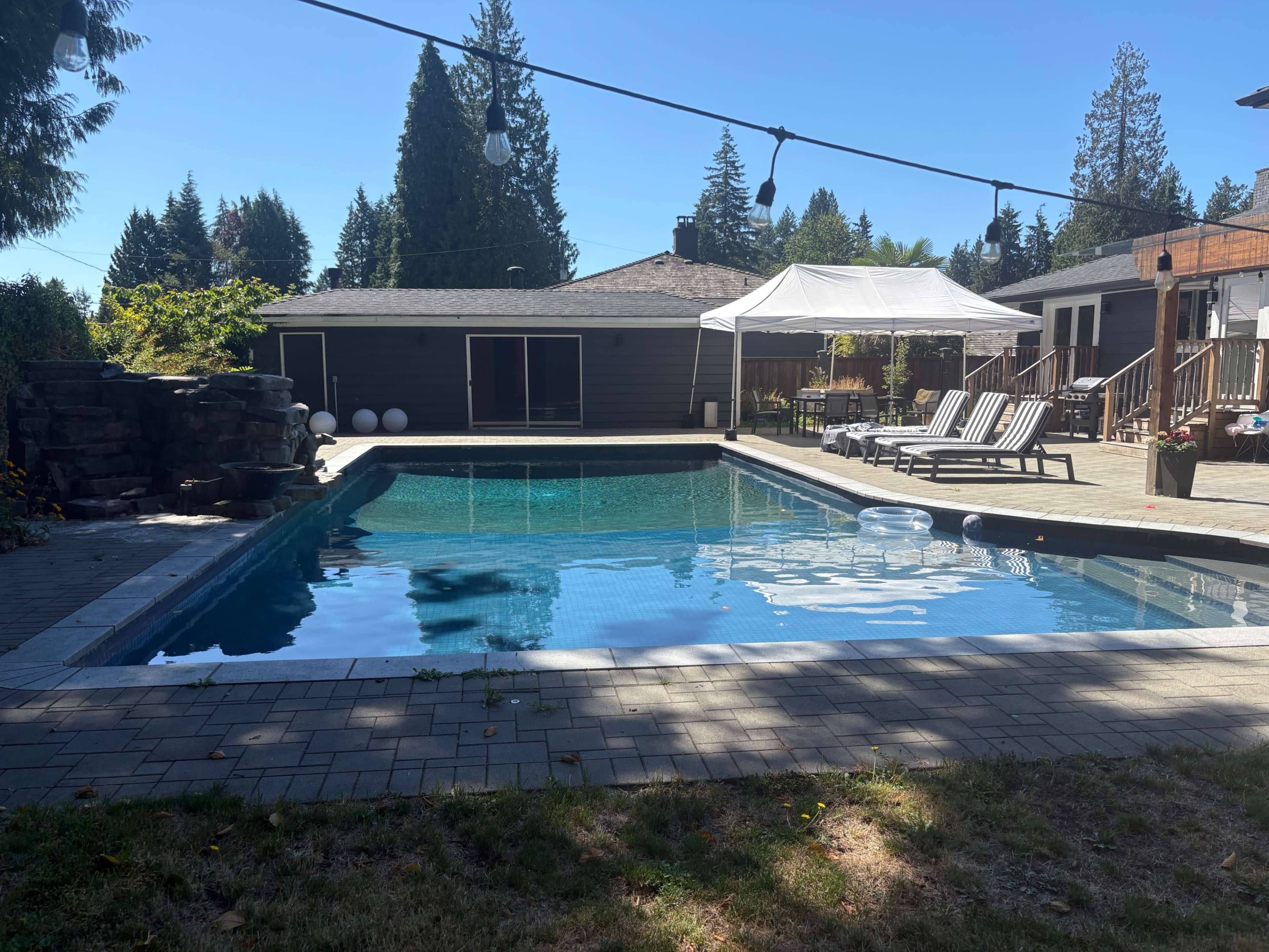 The image shows a backyard with a pool surrounded by lounge chairs, a gazebo, and a grassy area, set against a backdrop of trees and a clear blue sky.