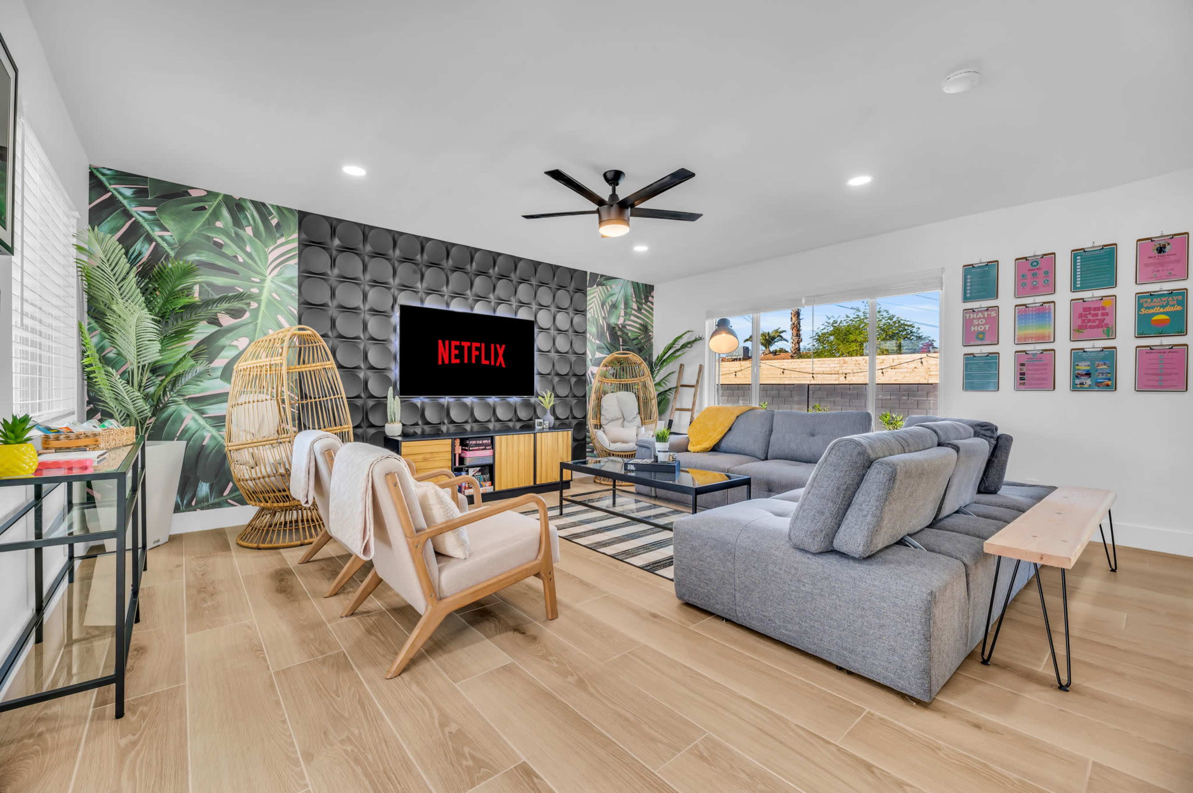 A modern living room features a large gray sectional sofa, two woven chairs, and a wall-mounted TV displaying the Netflix logo, set against a backdrop of tropical-themed wallpaper.