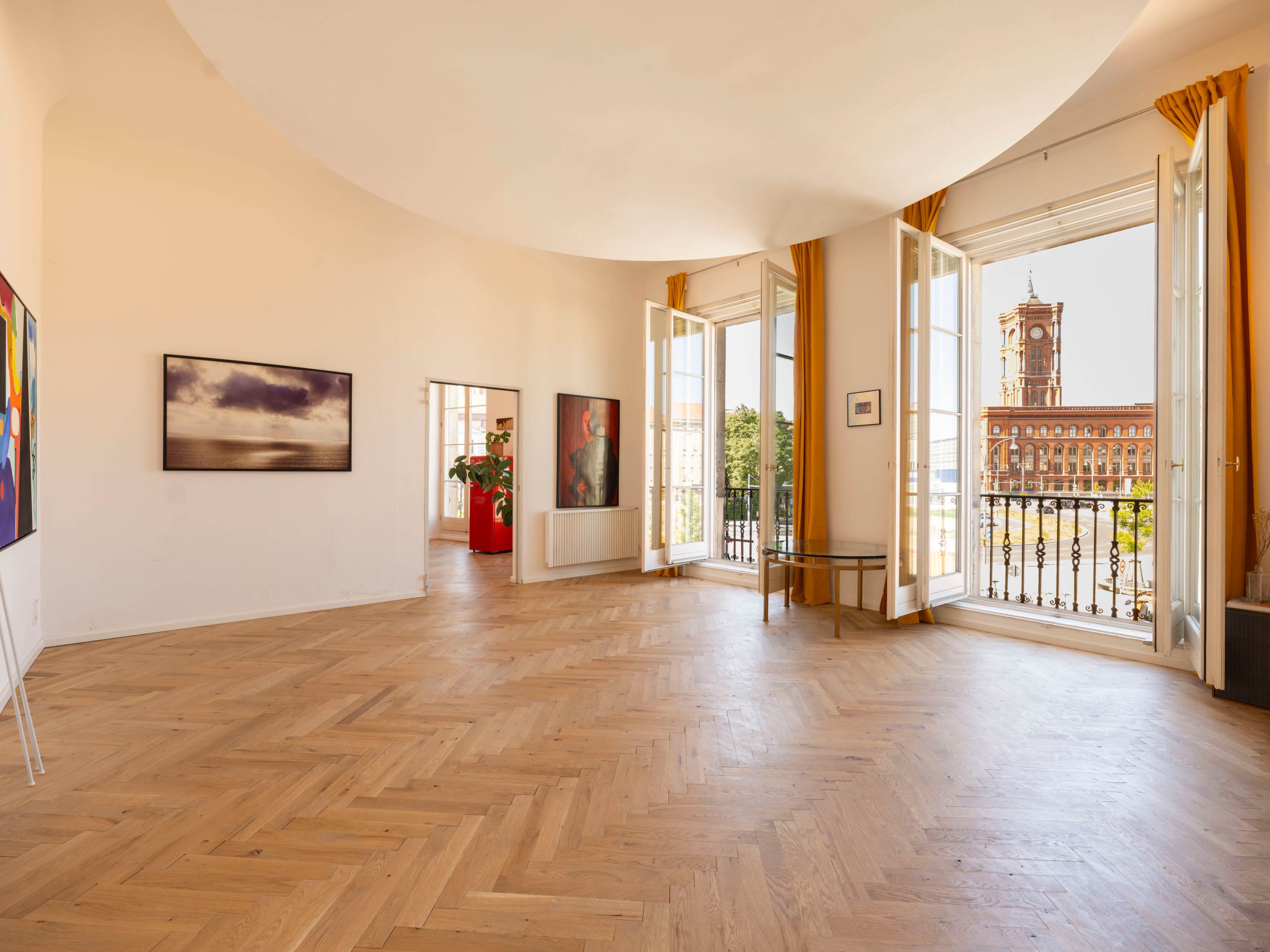 The image shows a spacious, well-lit room with large windows and wooden flooring, featuring artworks on the walls and a view of a building outside.