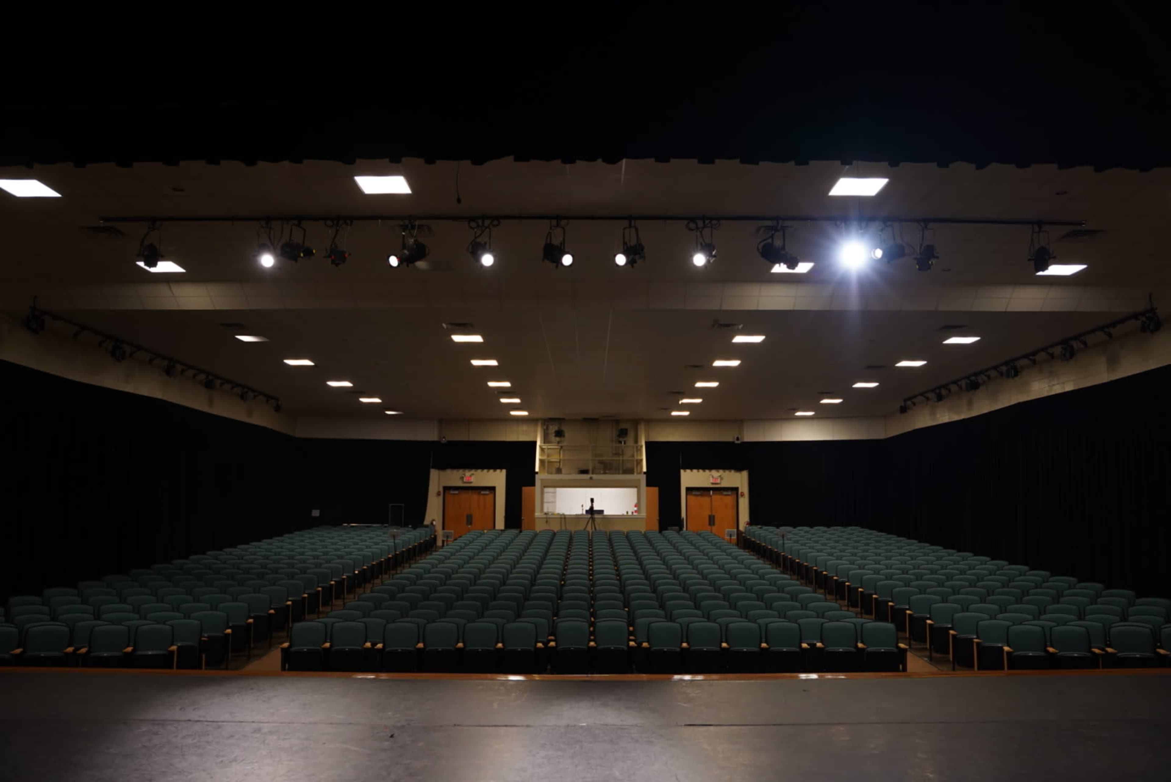 A conference hall with rows of green chairs facing a stage, illuminated by overhead lights.