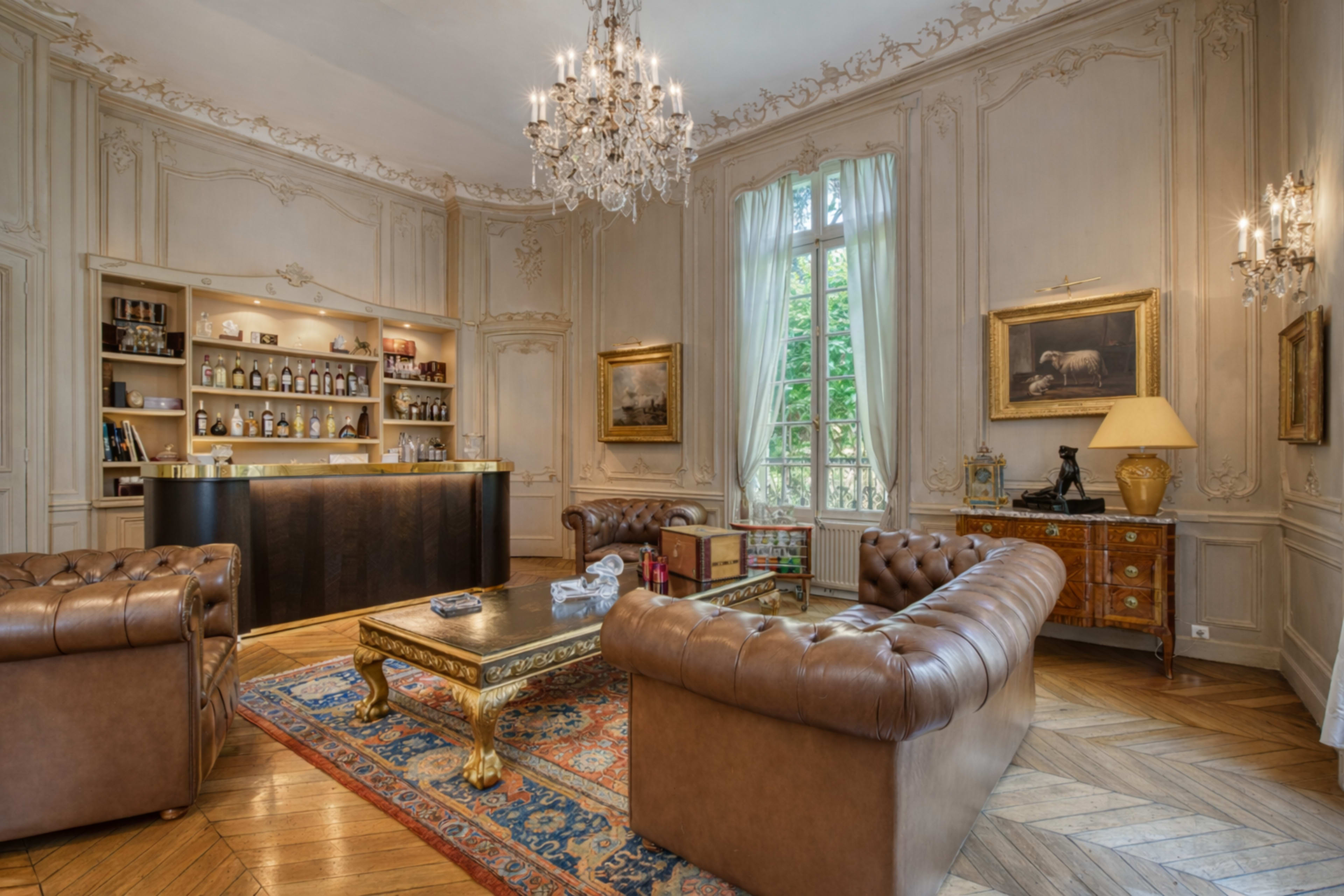 The image shows a luxurious room with two brown leather sofas, a wooden bar stocked with various bottles, and ornate decor featuring a chandelier and classical paintings.