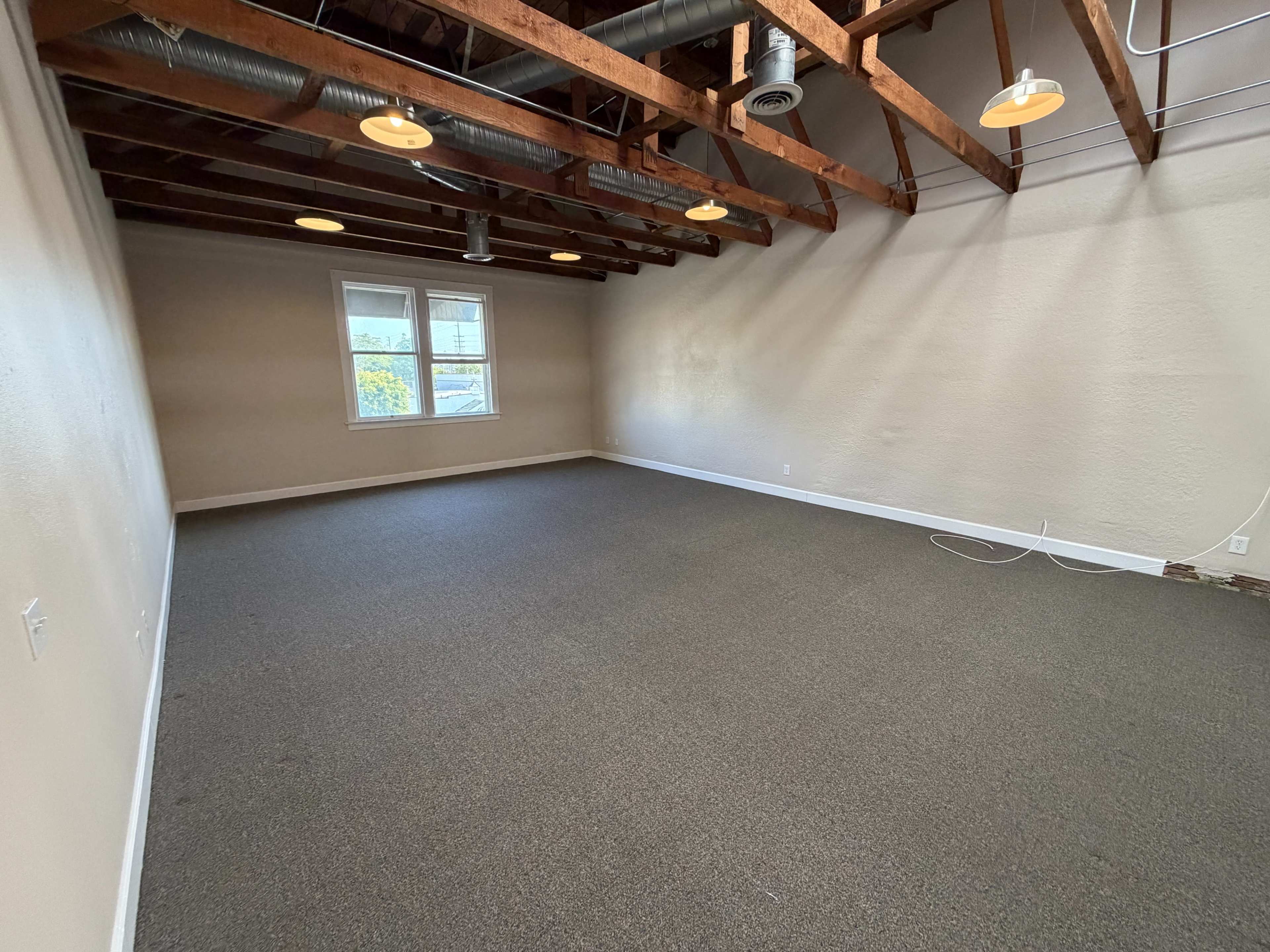 The image shows an empty room with exposed wooden beams, overhead lights, a carpeted floor, and a window along one wall.