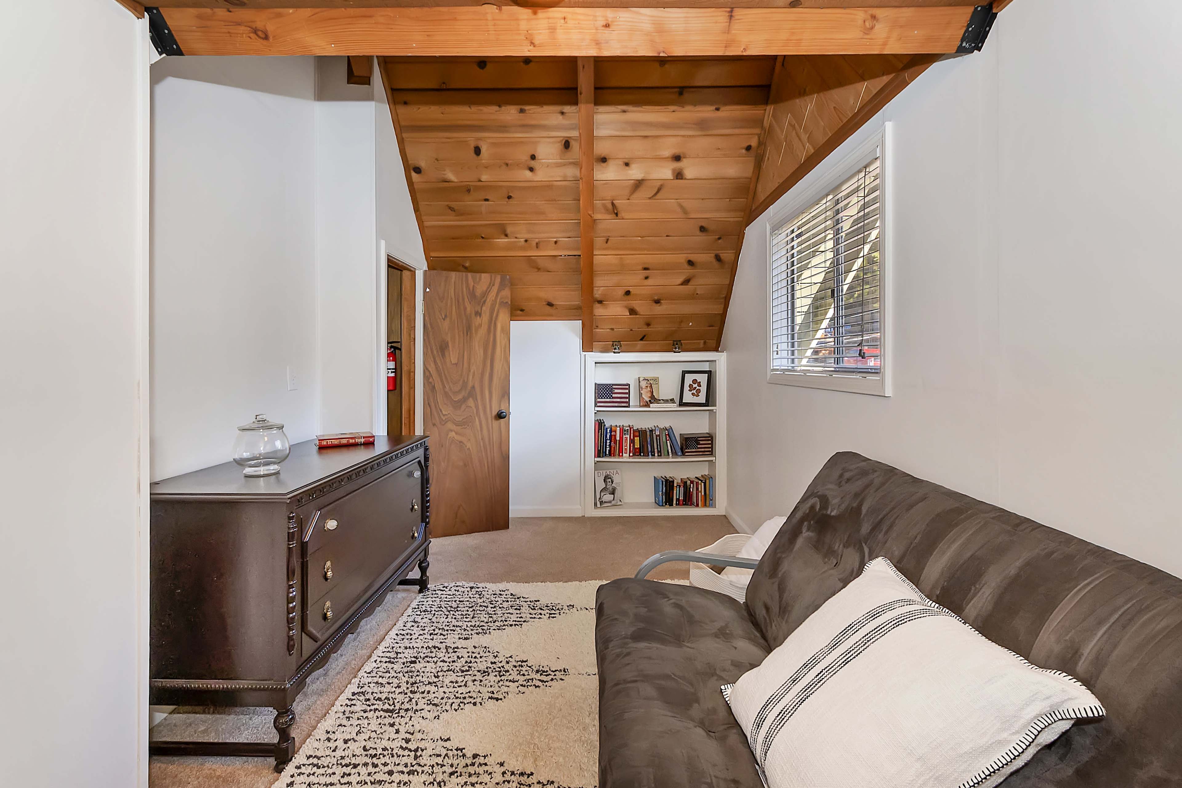The image shows a cozy room with a wooden ceiling, a dark dresser, a bookshelf filled with books, and a sofa with a decorative pillow, all set against neutral-colored walls.