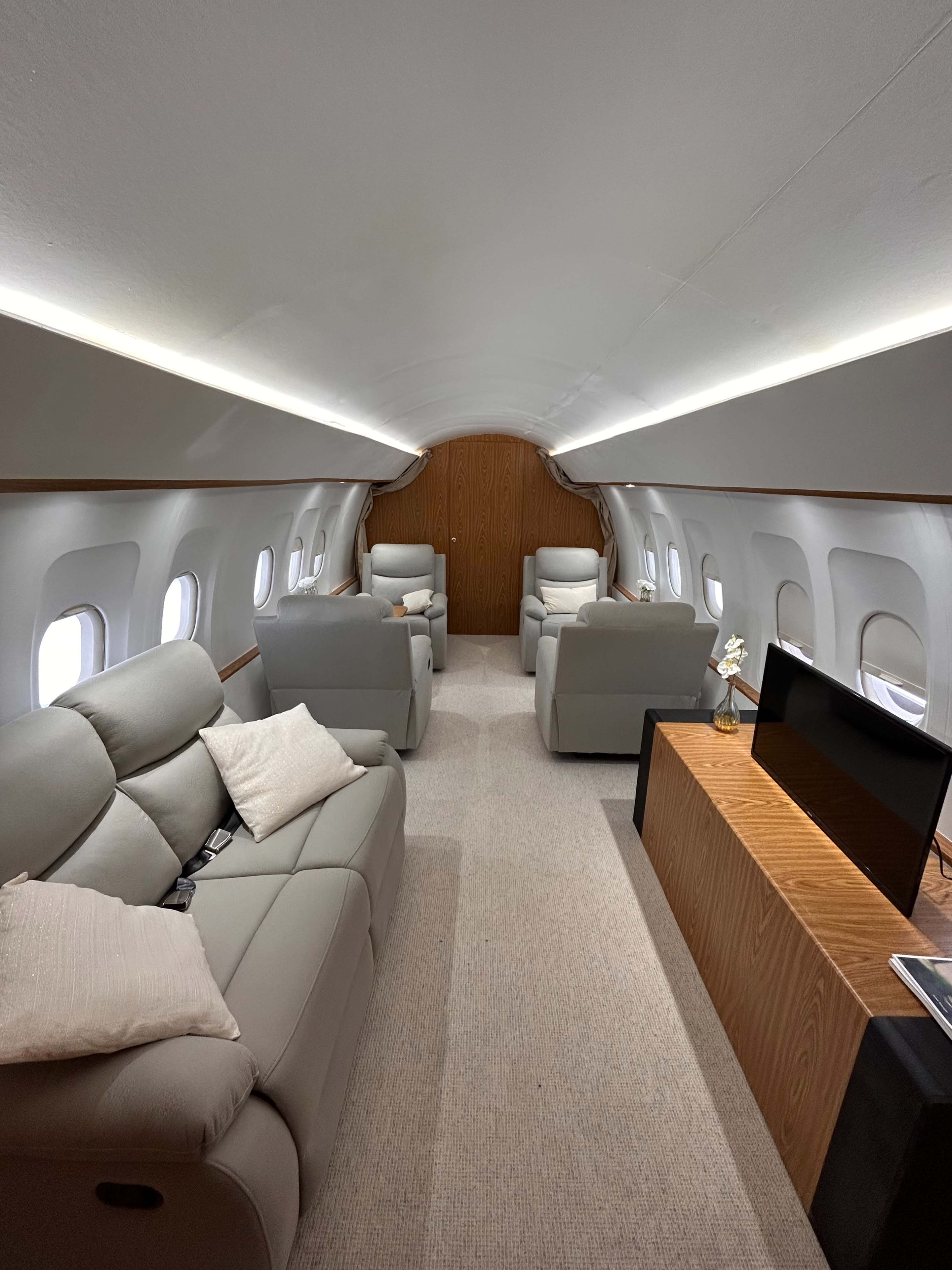 The interior of a private jet features two light gray sofas, several armchairs, and a wooden cabinet along the side, with large windows allowing natural light to illuminate the space.