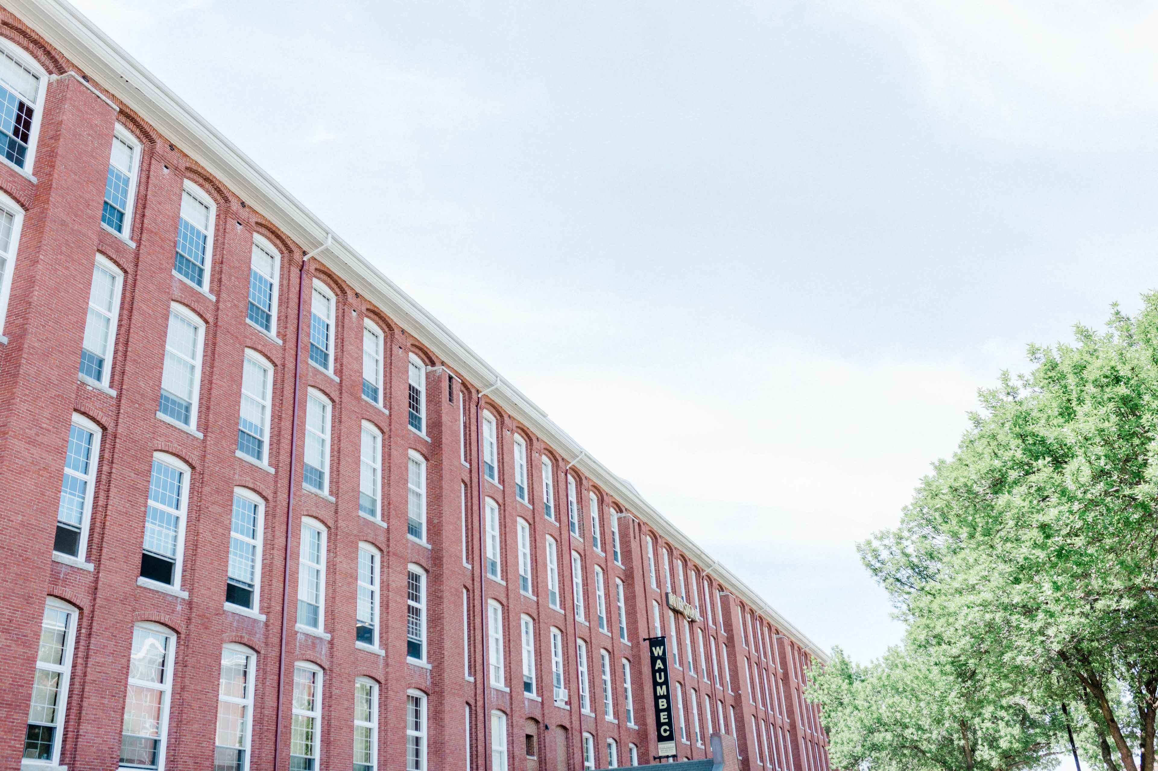 A long brick building with numerous windows, positioned against a clear sky and greenery on the right side.